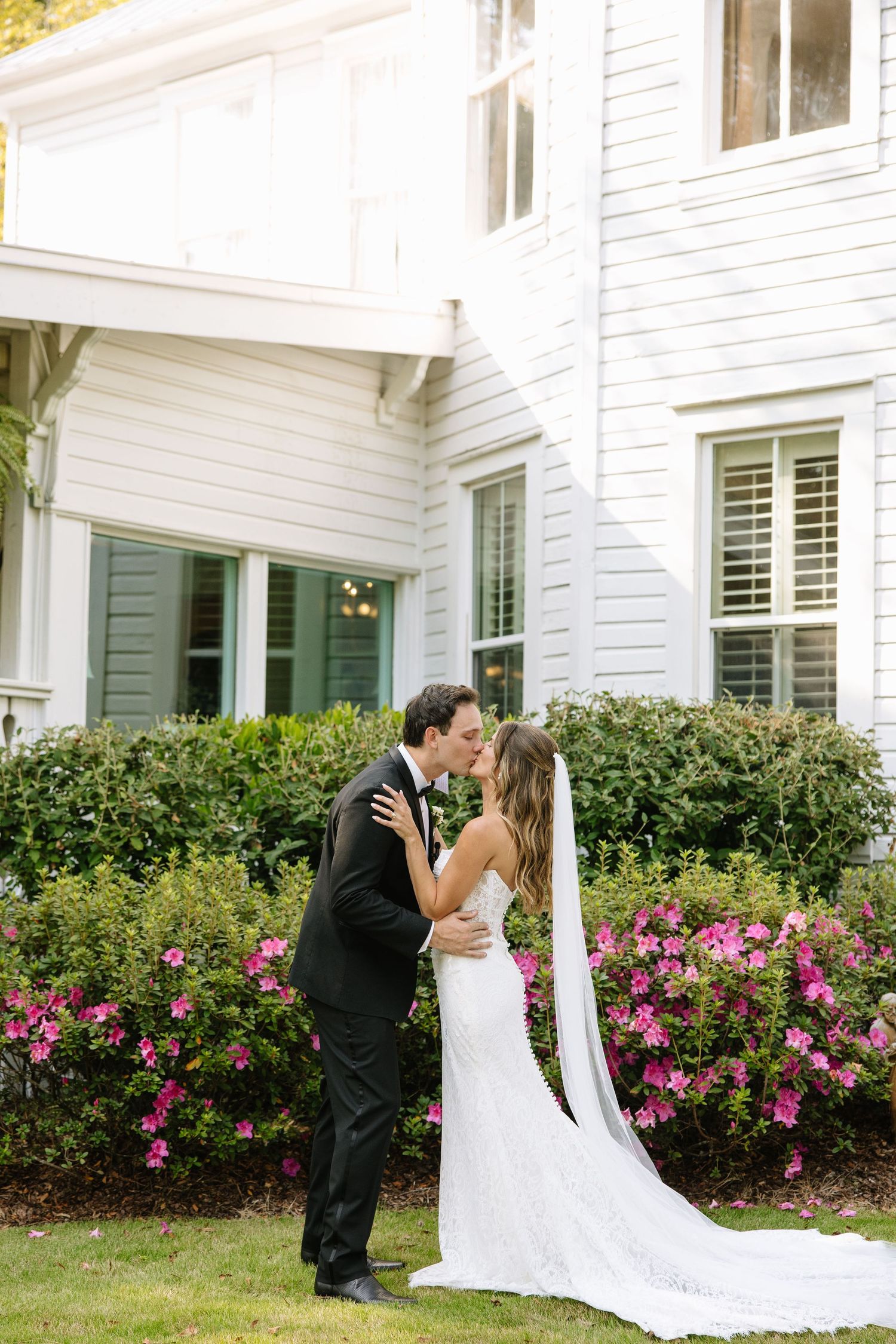 Wedding couple embracing in front of a classic white house with pink flower gardens.