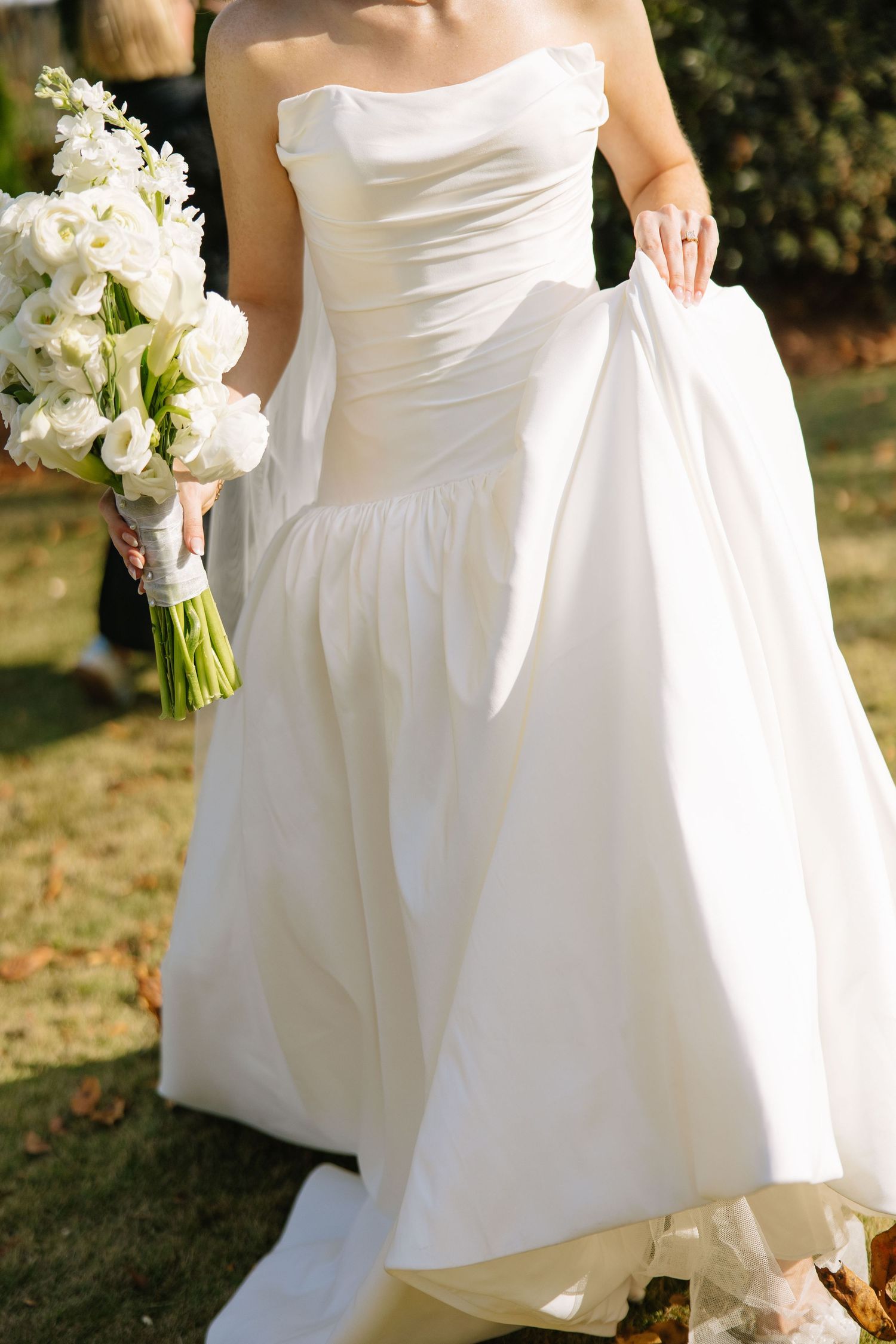 A bride in a flowing white wedding dress holds a bouquet of white flowers while standing on grass outdoors.