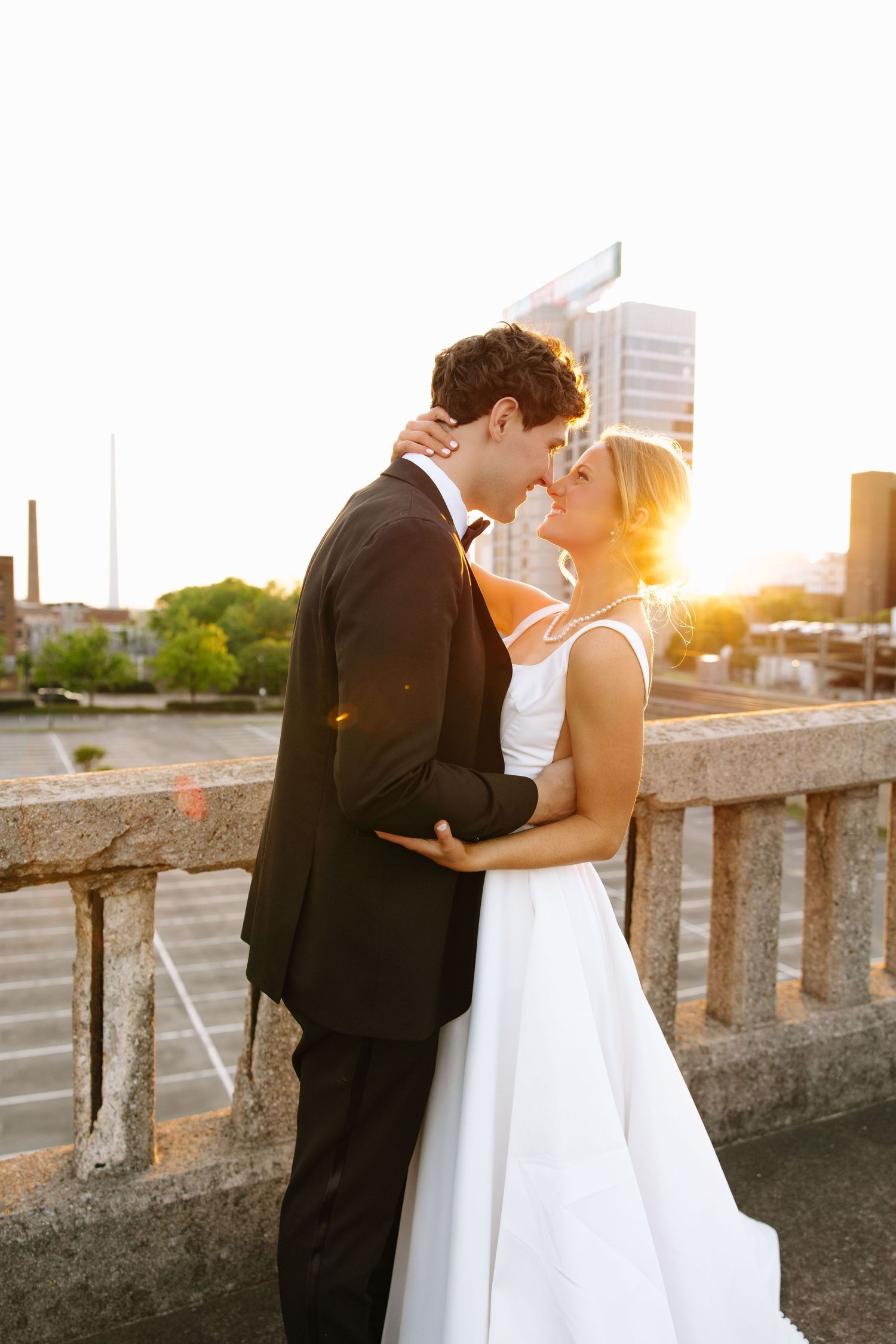 Bride and groom share a romantic sunset kiss on a bridge with city skyline in the background.