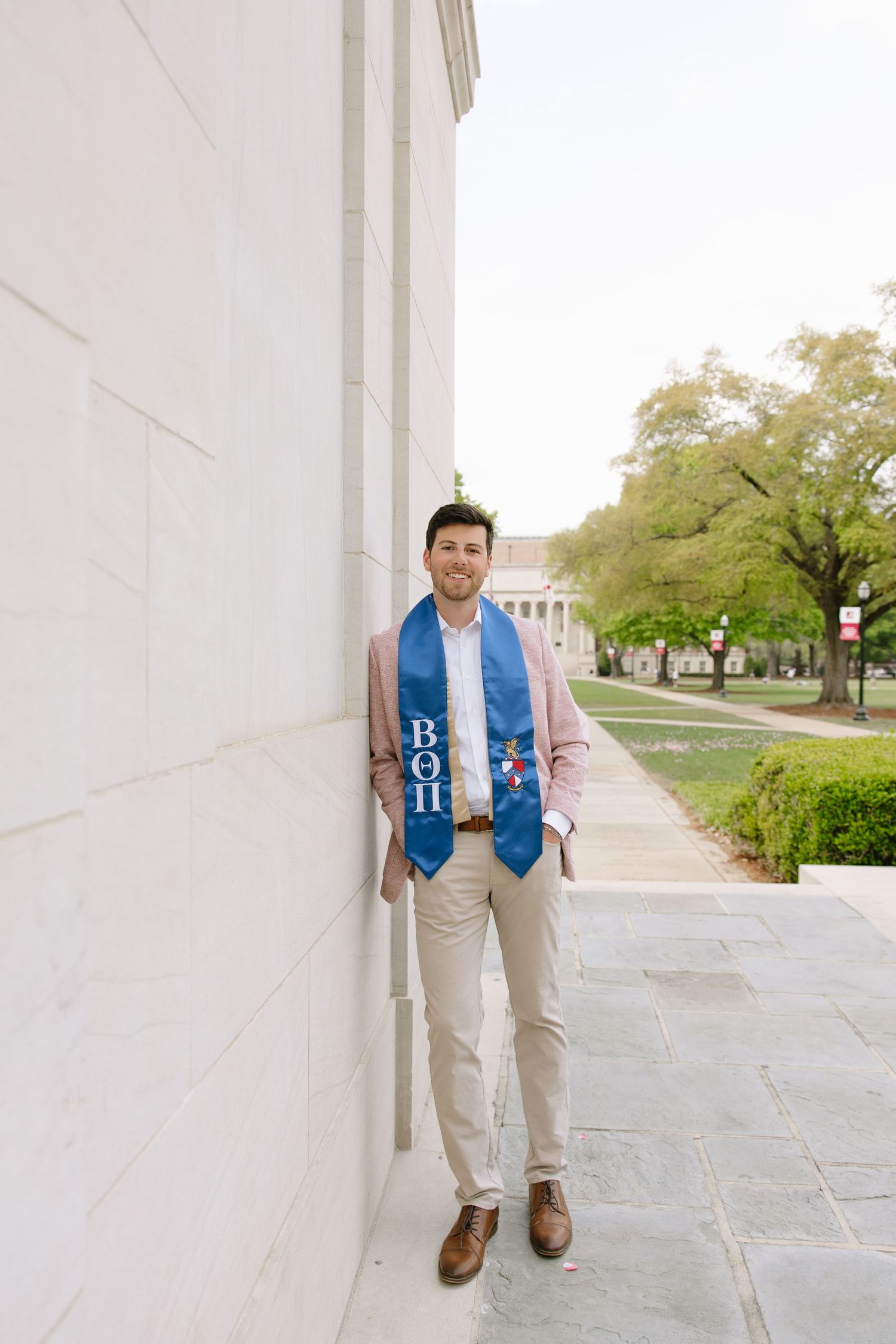 College graduate smiling in blue fraternity stole and blazer, leaning against white column on campus walkway.