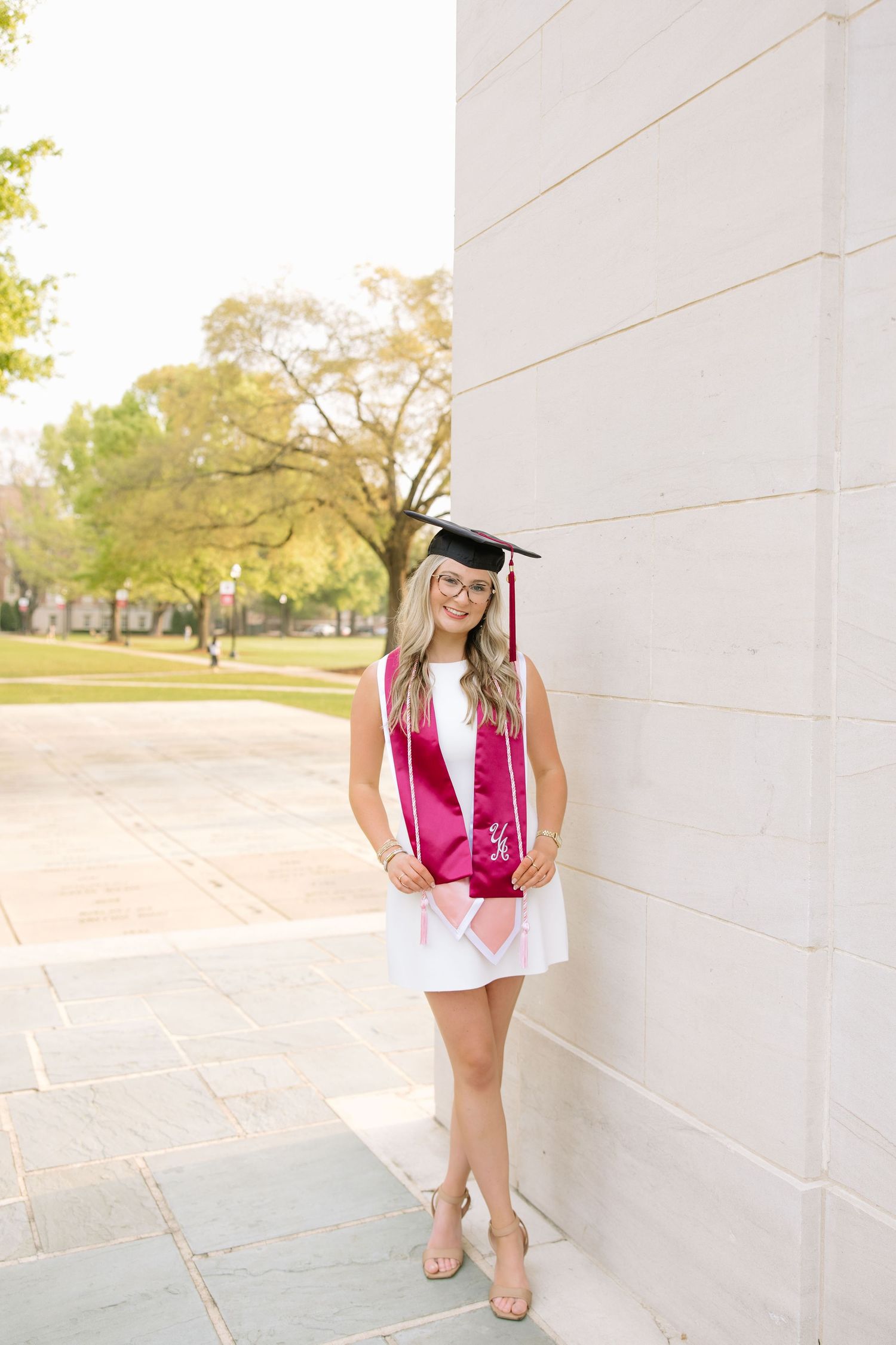 Smiling female graduate in white dress, maroon cap and gown stole, poses against stone column on sunny college campus.