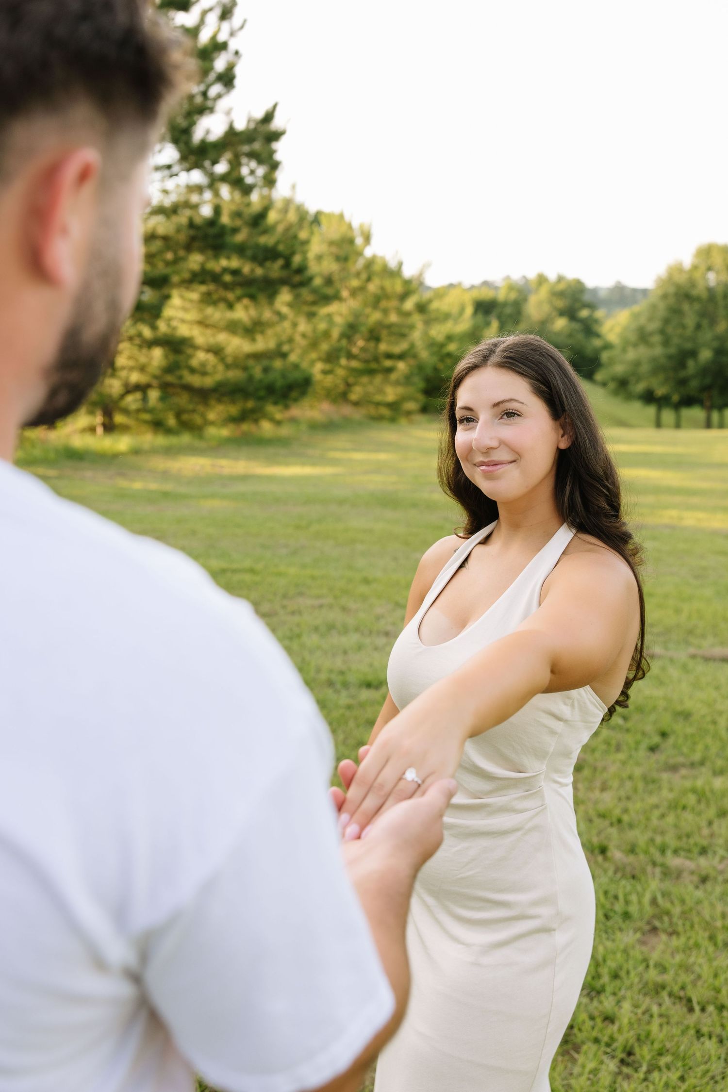 A smiling brunette woman in a white dress holds hands with a man outdoors in a green park, showing off an engagement ring.