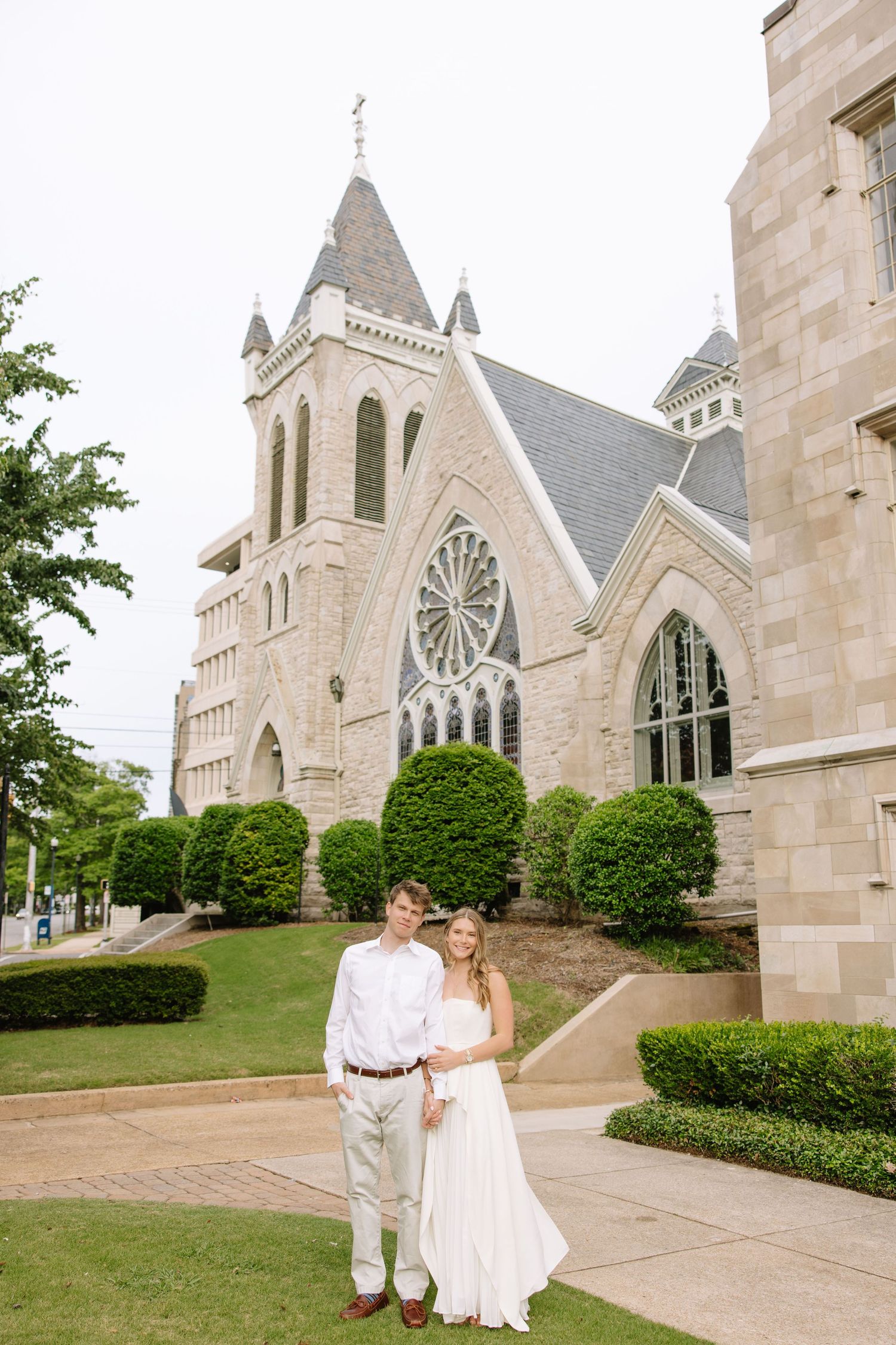 Couple in white attire poses before a Gothic stone church with rose window and manicured hedges.