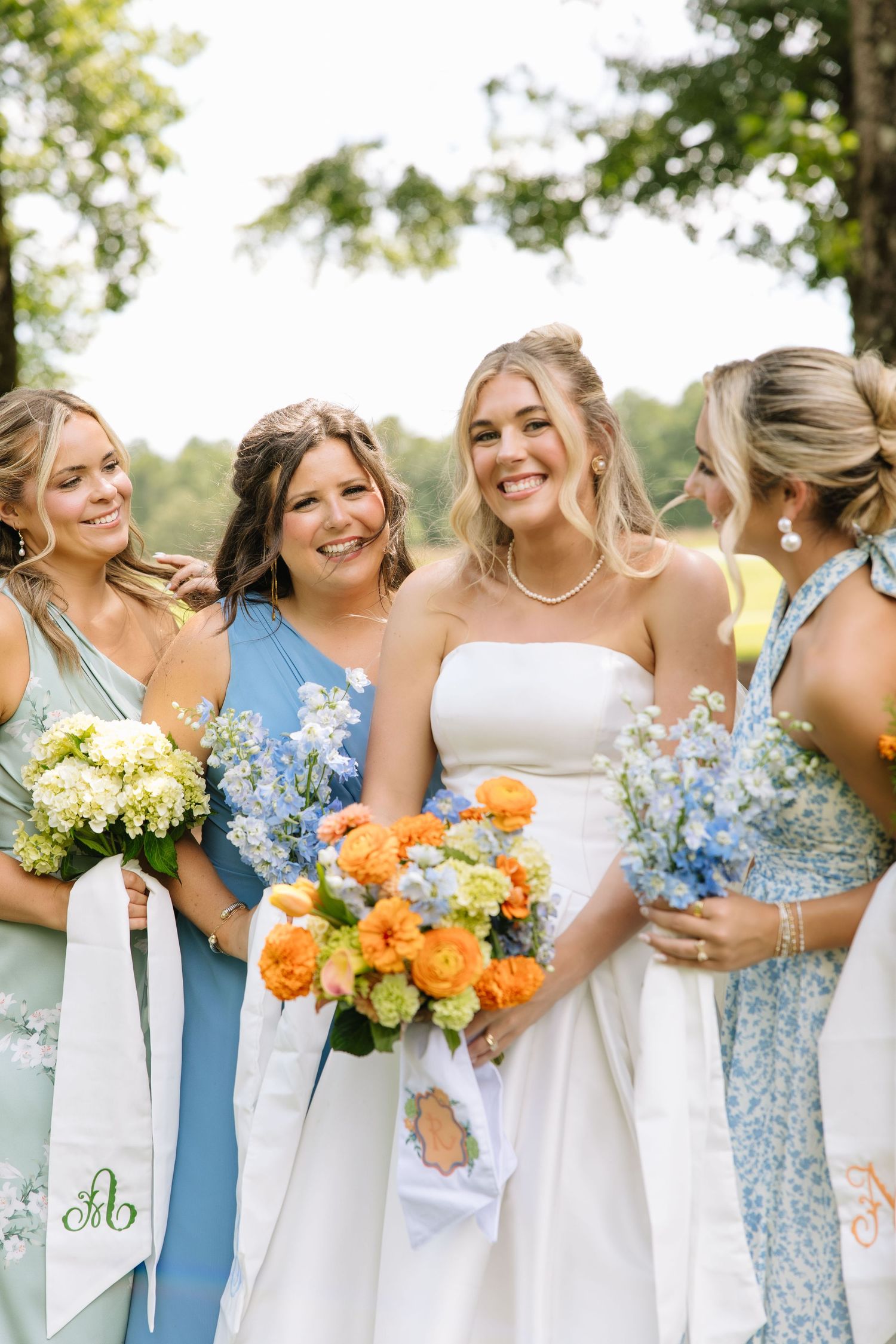 Bride in white strapless gown holds orange bouquet surrounded by bridesmaids in blue dresses with floral bouquets outdoors.
