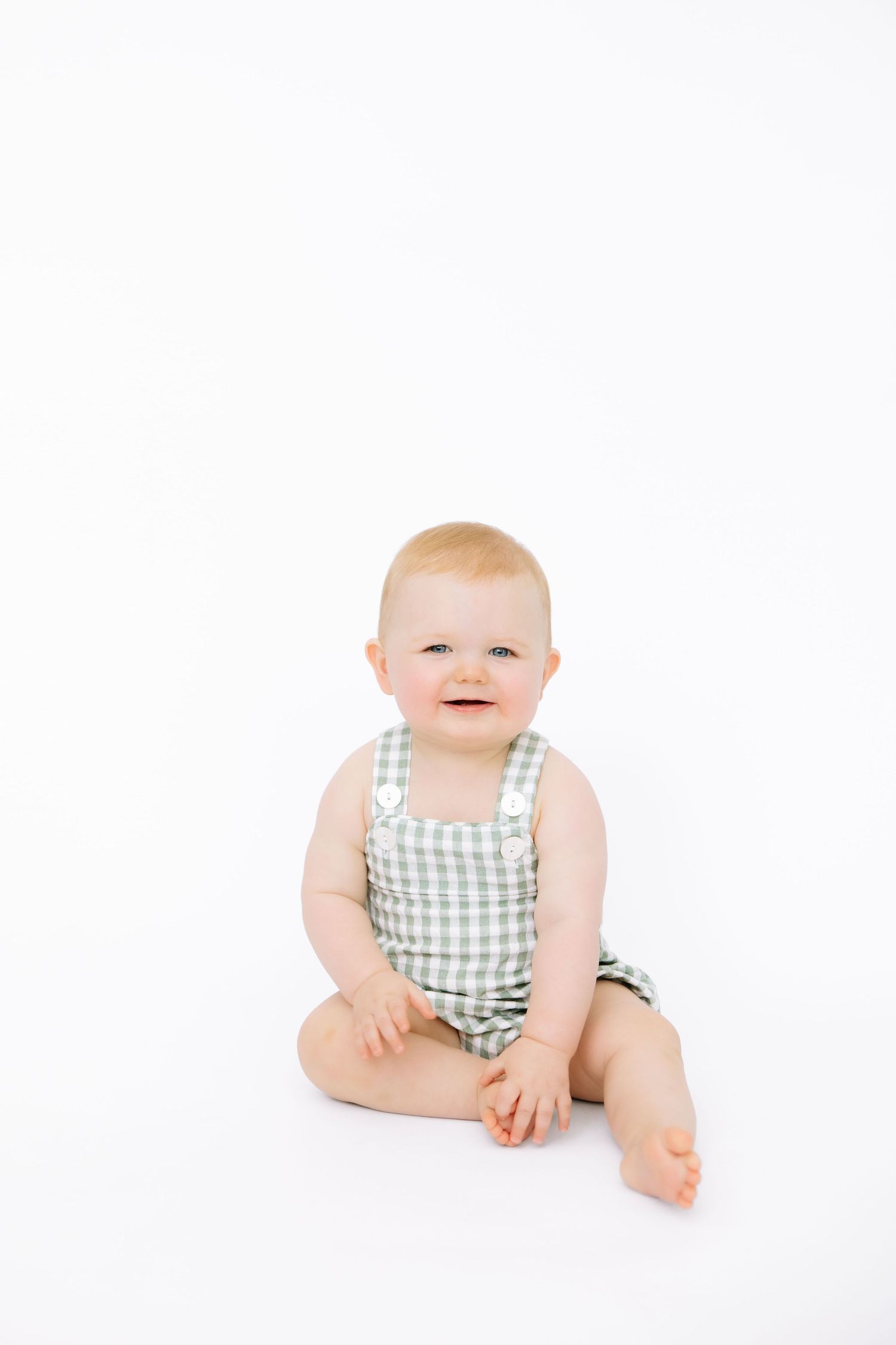 Smiling baby in green gingham overalls sitting on white background.
