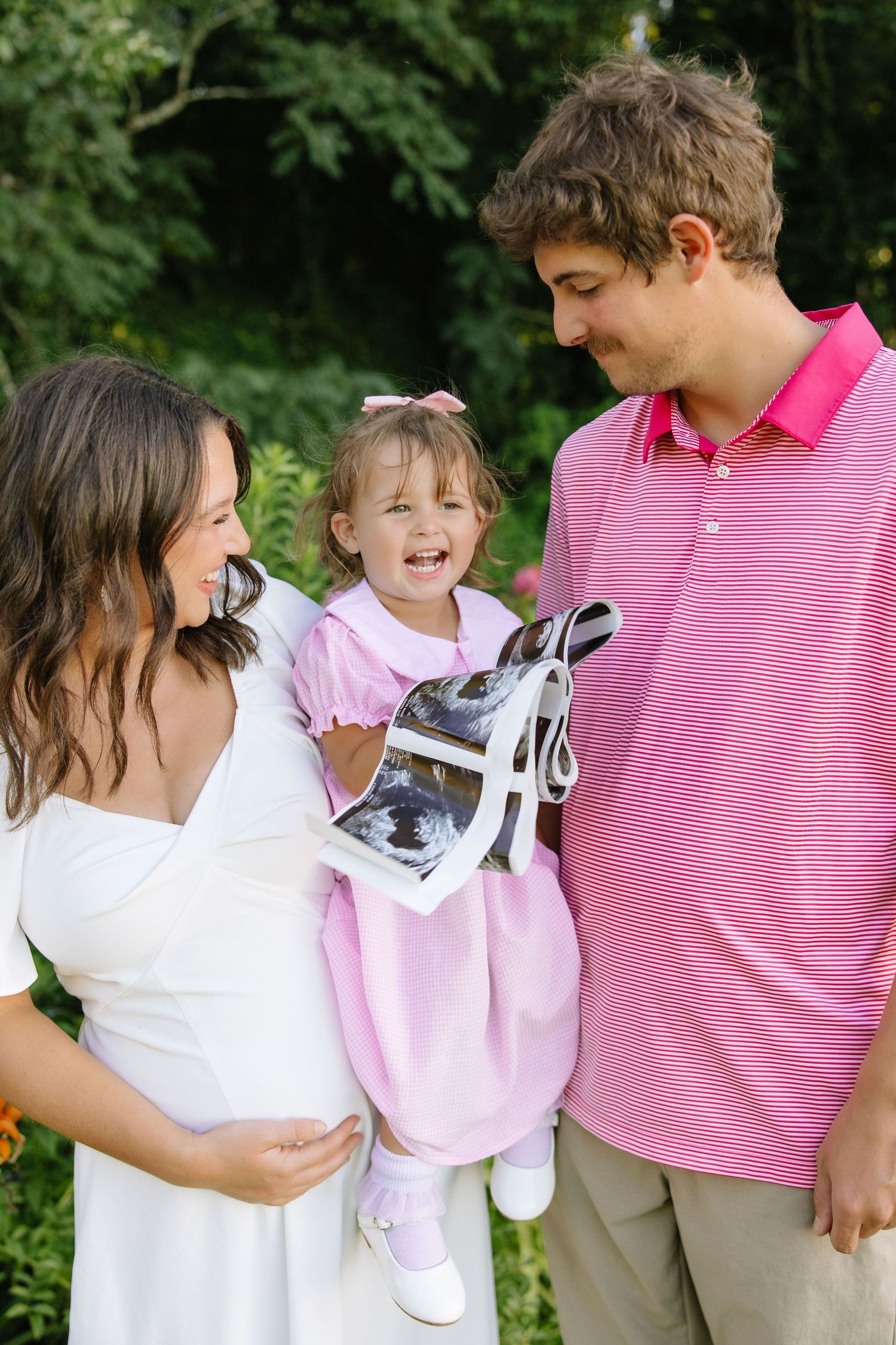 Happy family with pregnant mother, toddler girl holding ultrasound photo, and father smiling outdoors.