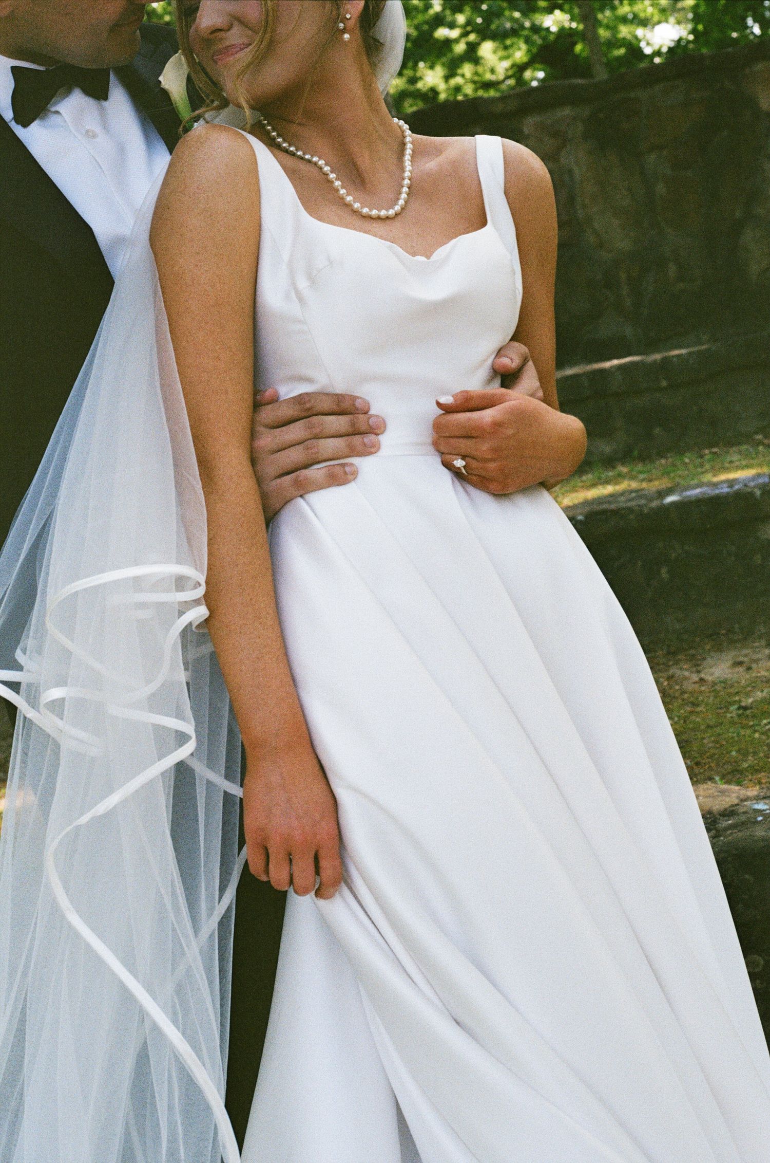 Close-up of bride in elegant white gown with pearl necklace and veil standing with groom outdoors.