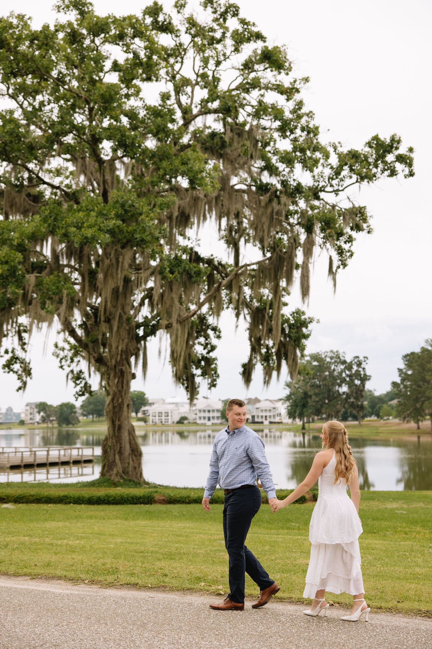 Couple holding hands walking near a scenic lake with Spanish moss-draped oak tree at an outdoor engagement session.