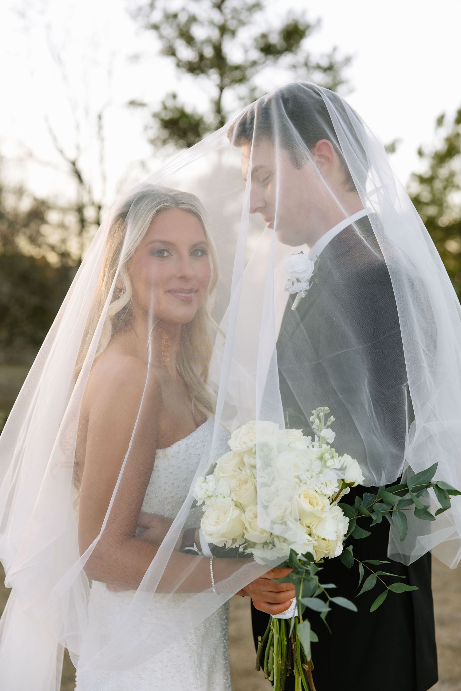 Romantic portrait of couple under flowing veil with white bouquet.