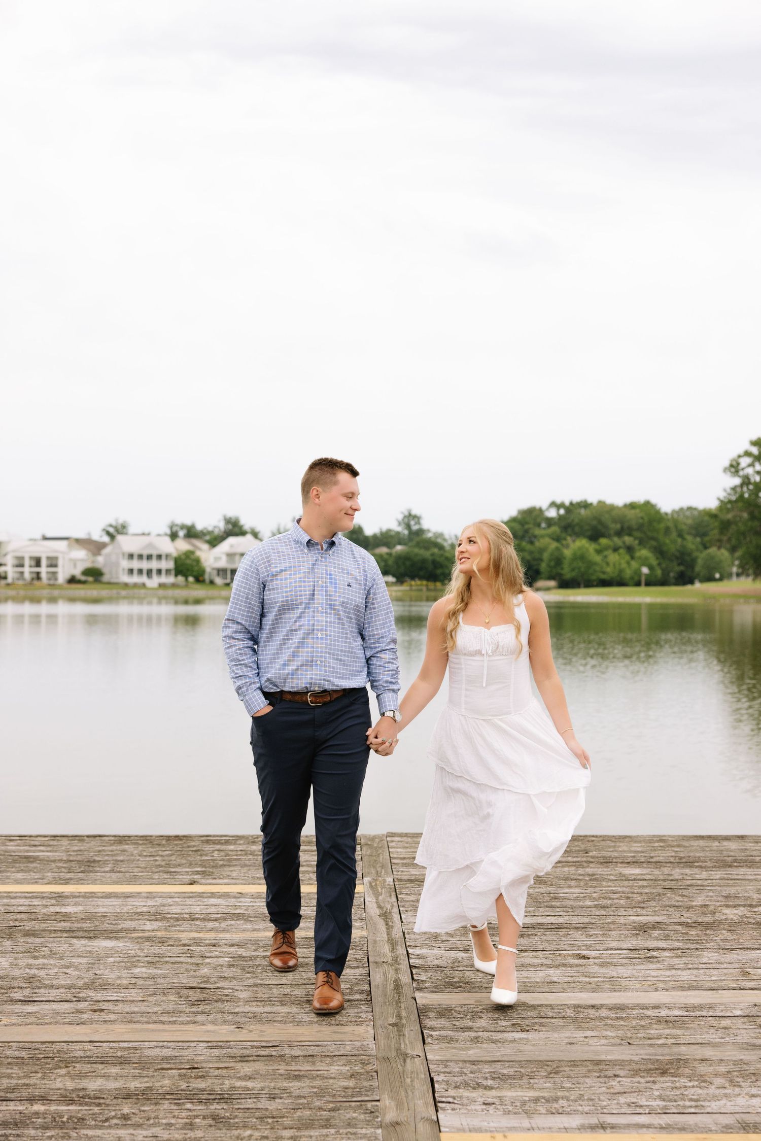 A couple holds hands and walks on a wooden dock by a lake, the woman in a white dress and the man in a blue shirt.