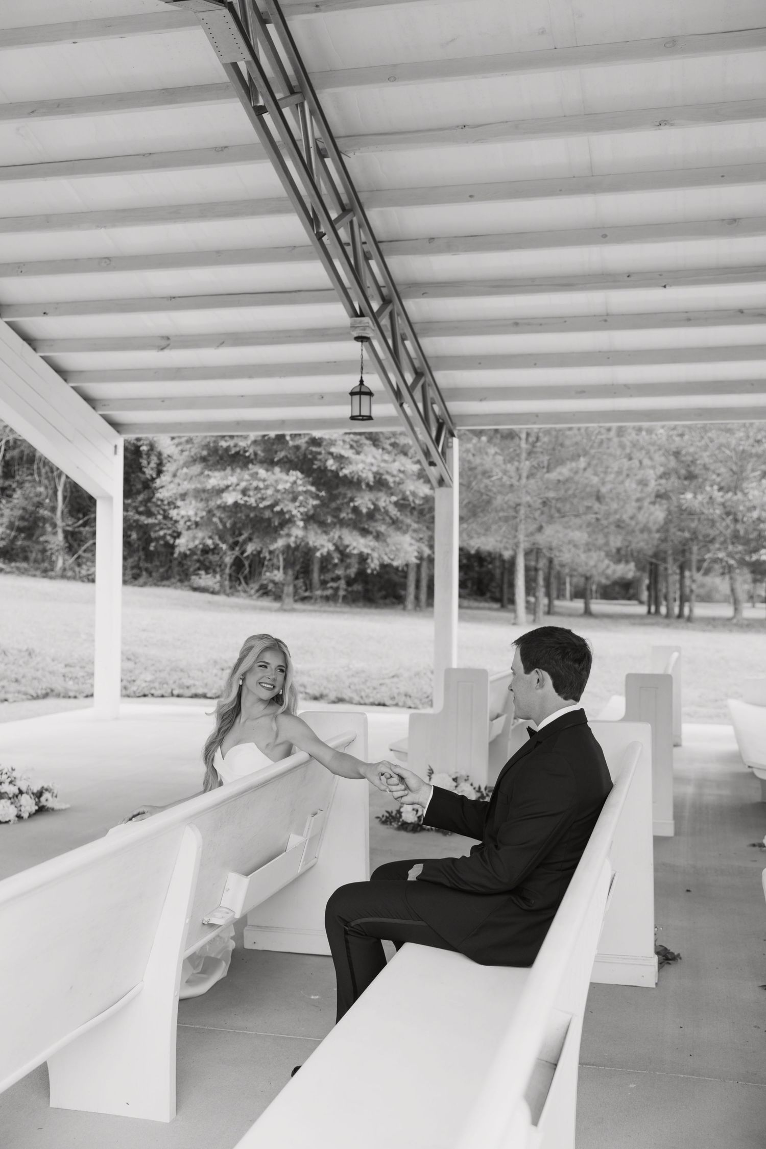 Black and white photo of bride and groom holding hands on white ceremony benches under an elegant outdoor pavilion.