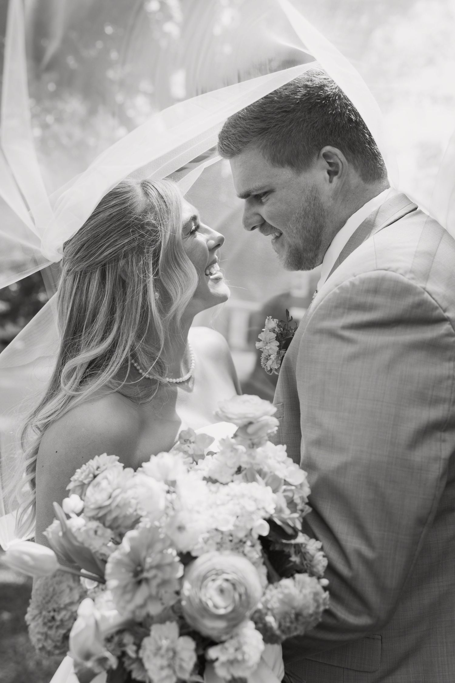 Black and white photo of a smiling bride and groom under a veil, bride holding a floral bouquet.