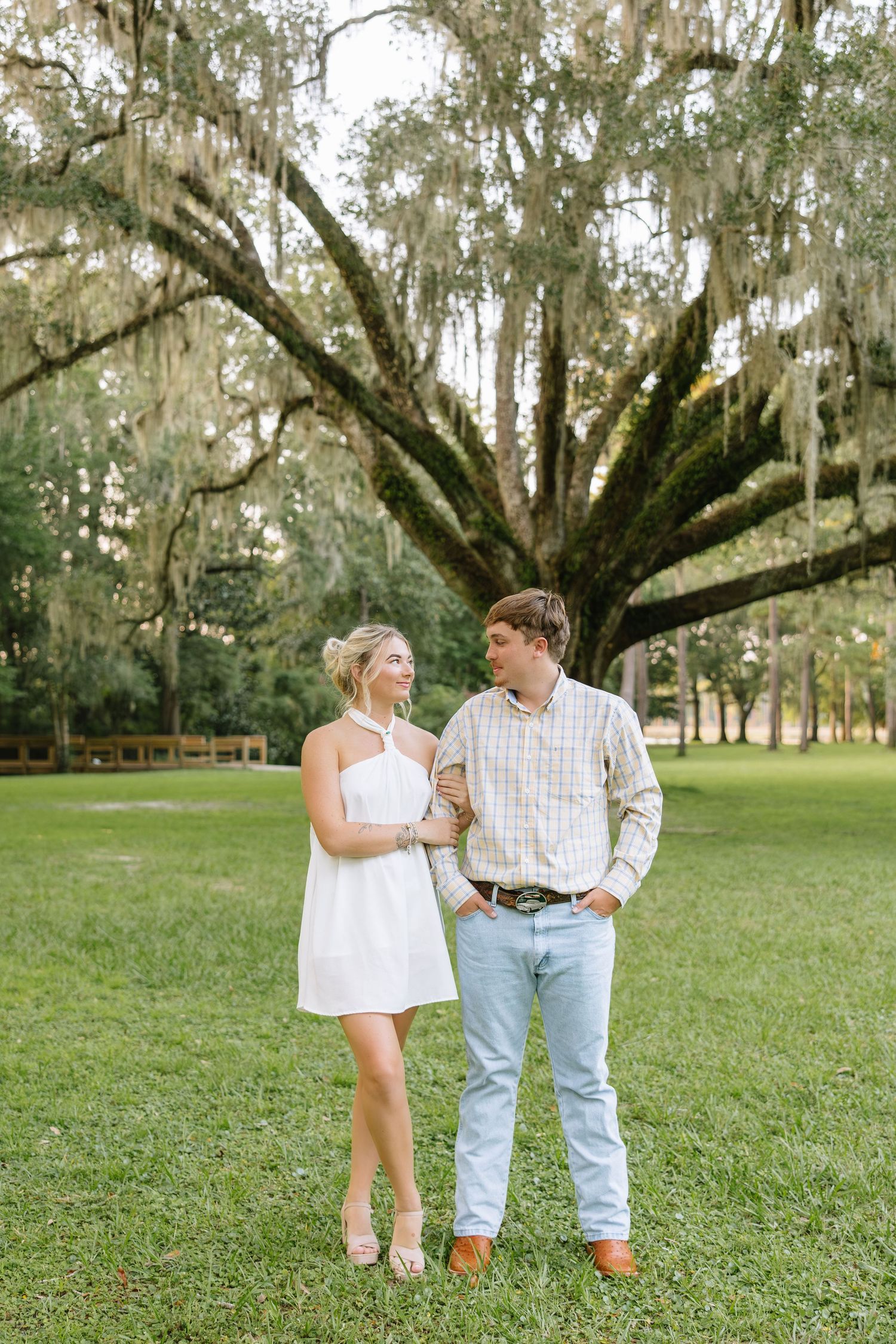 Couple standing together on green grass beneath a large oak tree draped with Spanish moss.