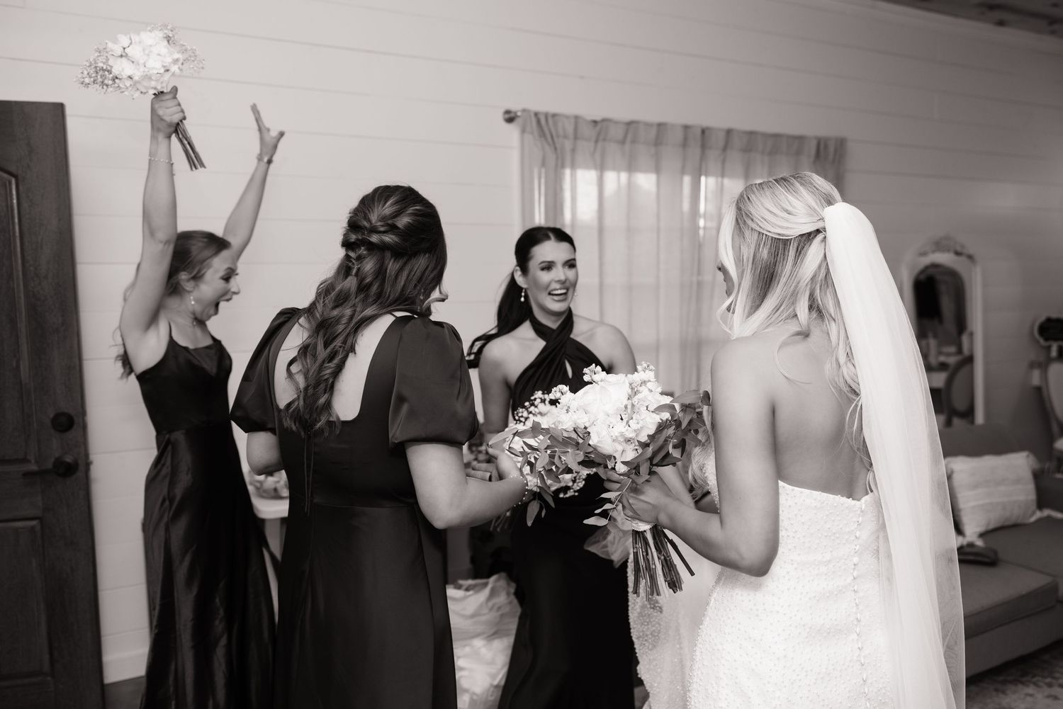 Bridesmaids in black dresses celebrate with bouquets in bridal suite.