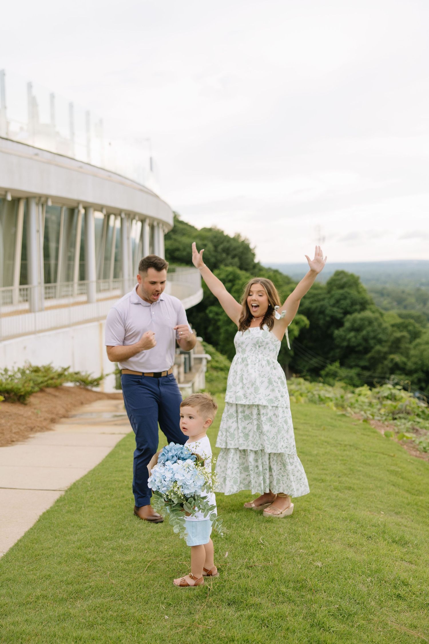 A joyful family of three celebrates outdoors, woman in floral dress raises arms, boy holds blue hydrangeas, elegant venue behind them.