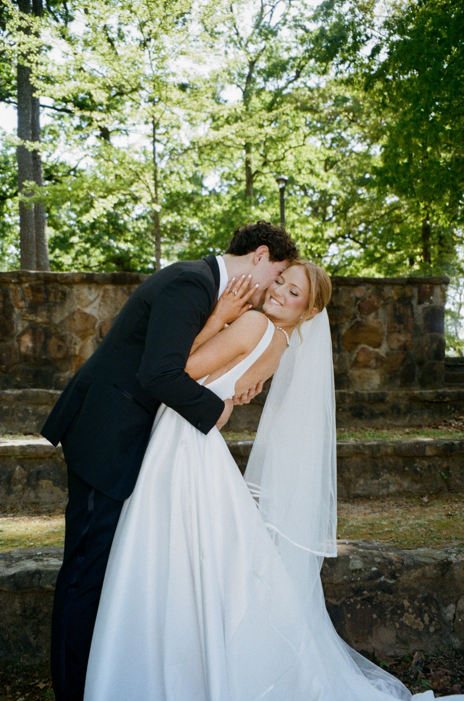 Groom tenderly kisses bride on cheek outdoors near stone wall, bride smiling in white gown and veil.