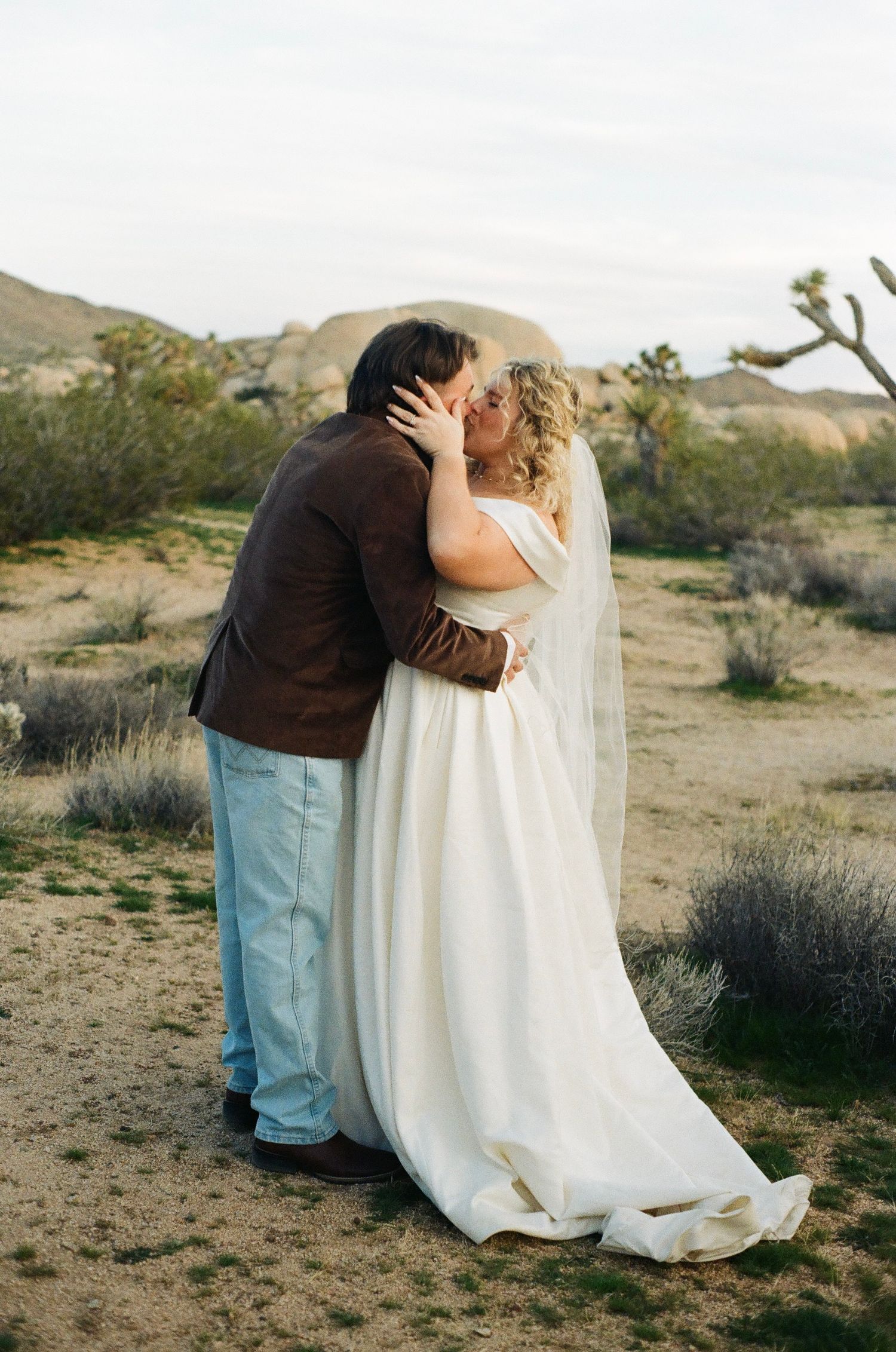 A couple sits together in a desert landscape with scrub brush and mountains in the background.
