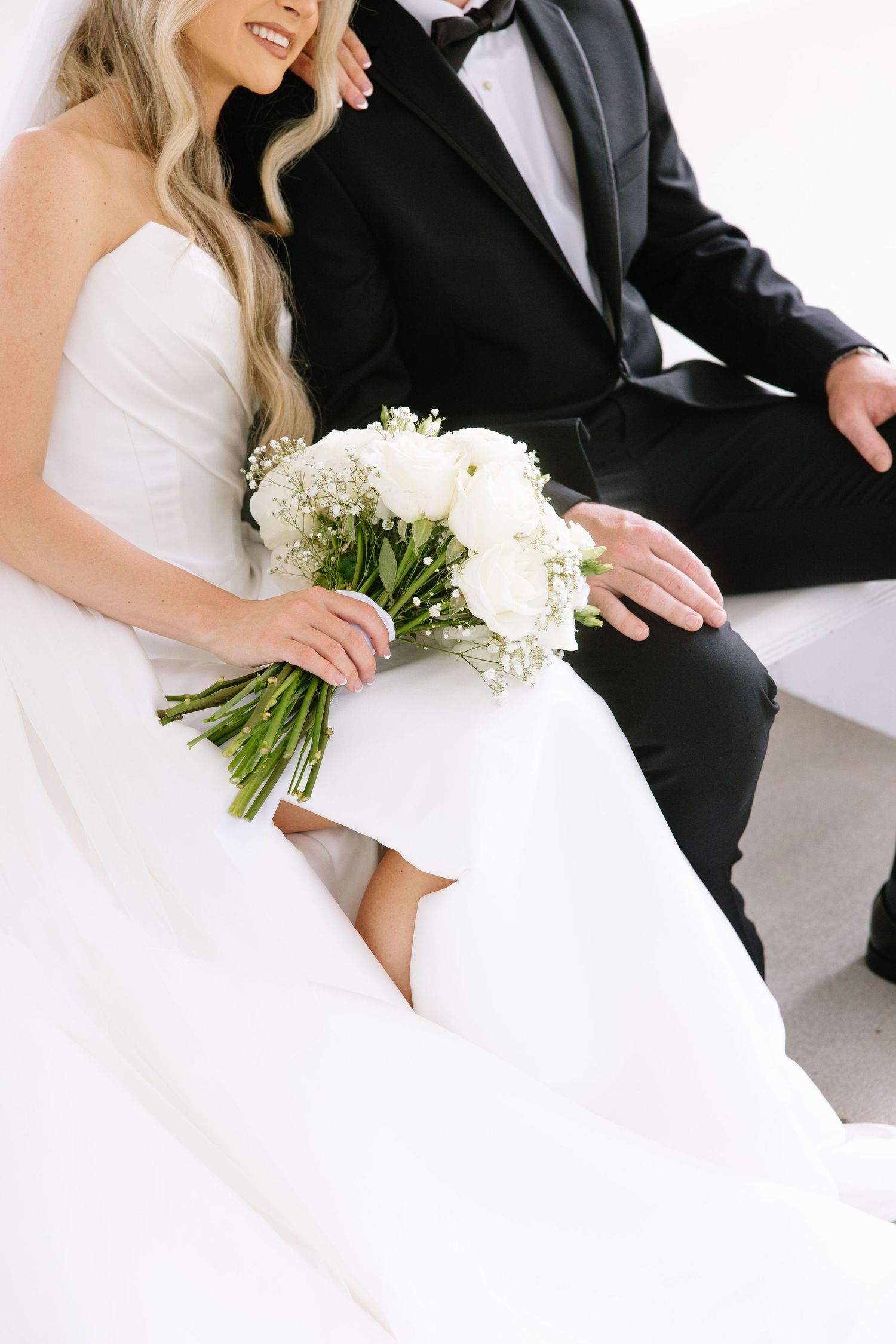 Bride in white gown holding bouquet of white flowers sits beside groom in black tuxedo at wedding ceremony.