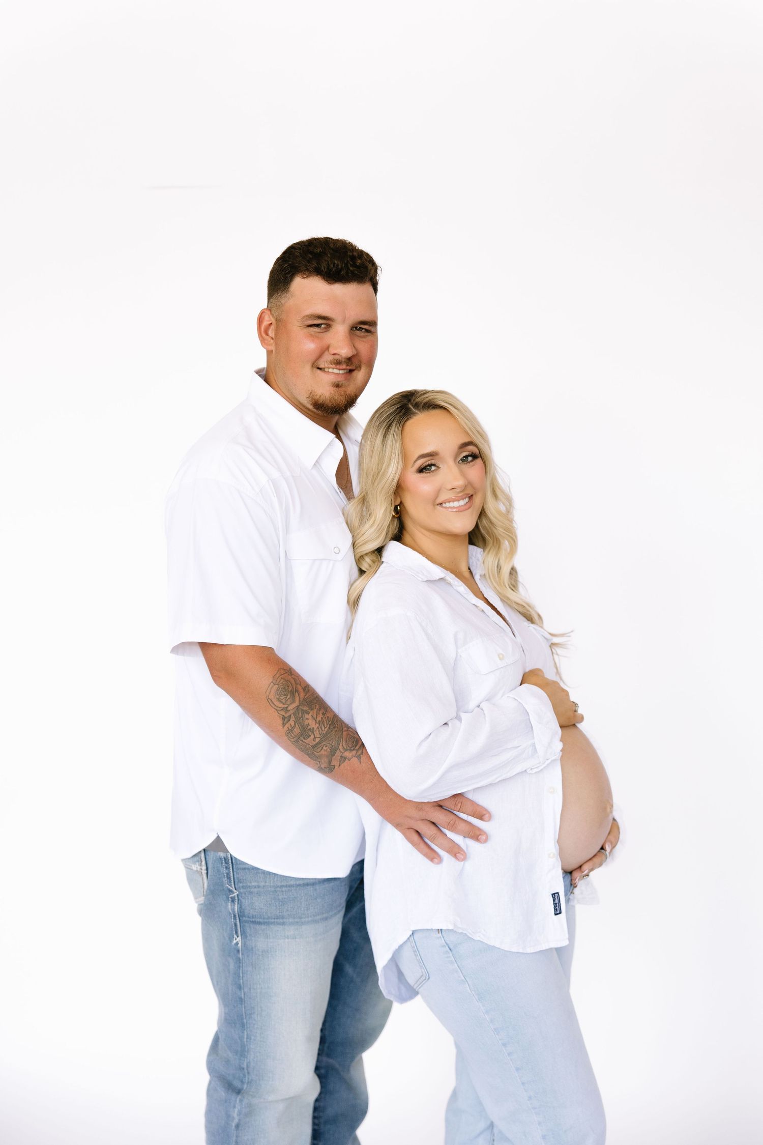 A smiling couple in matching white shirts and jeans poses for a maternity photo, with the woman cradling her baby bump.