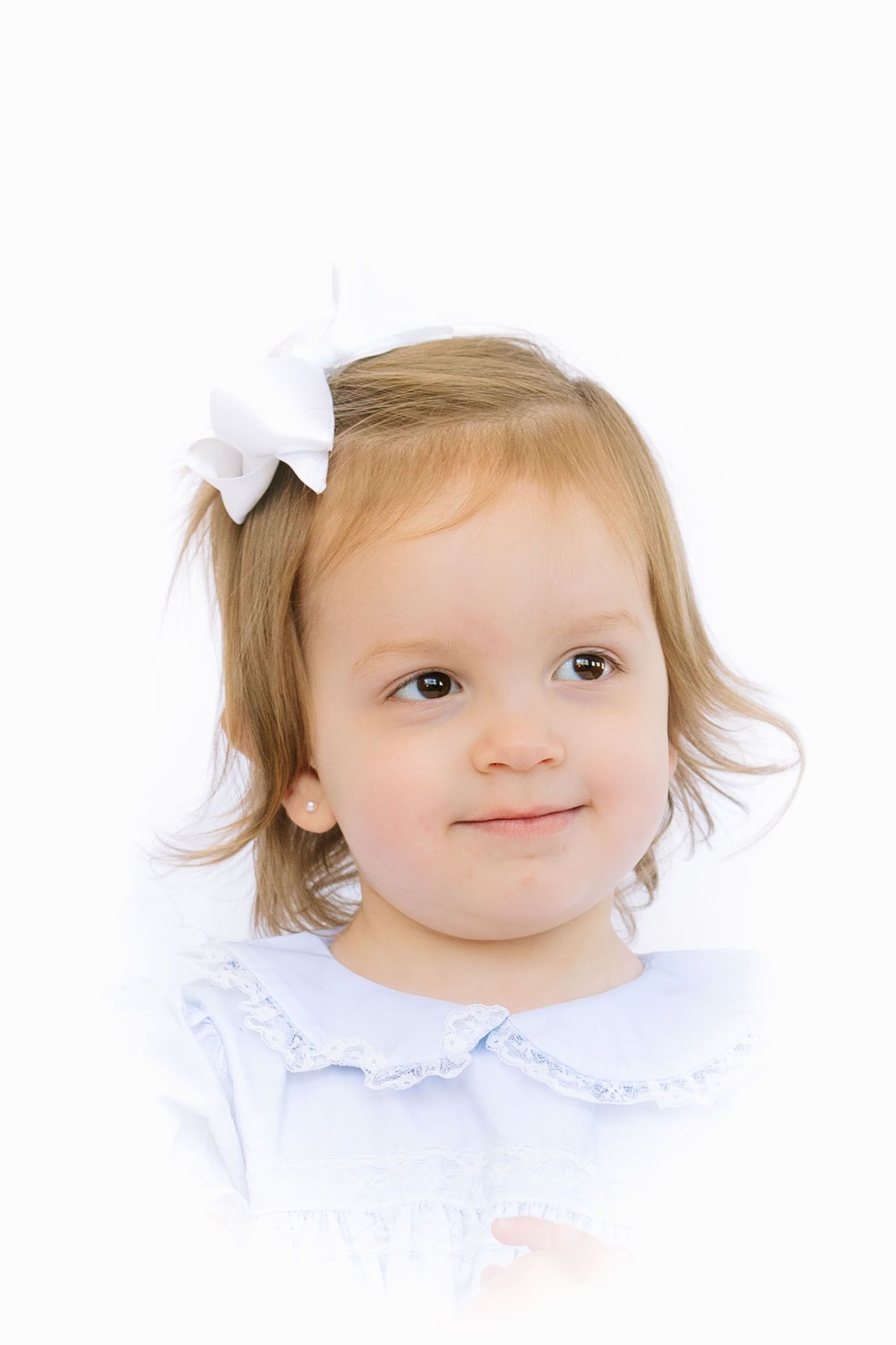 Adorable toddler girl with a white bow in her hair wearing a light blue dress, smiling against a white background.