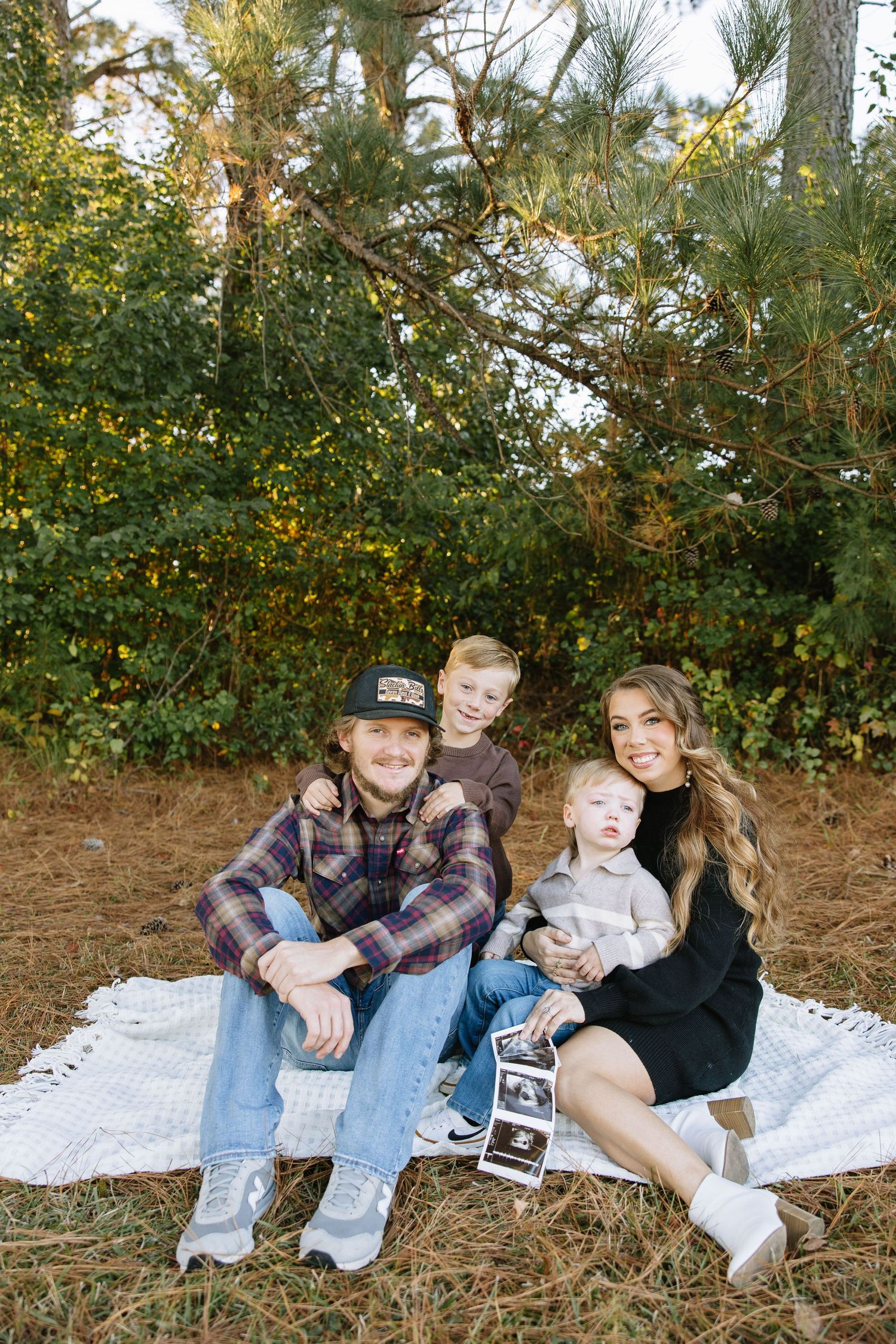 Family of four sits on blanket outdoors holding ultrasound photos, announcing pregnancy among pine trees.