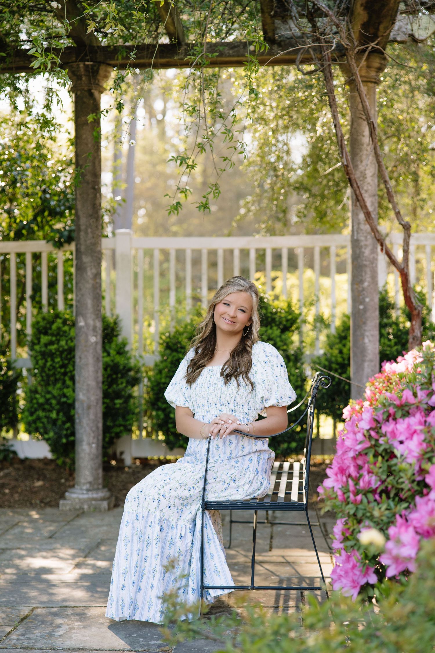 Young woman in floral dress sitting on garden bench under pergola surrounded by pink azaleas and lush greenery.
