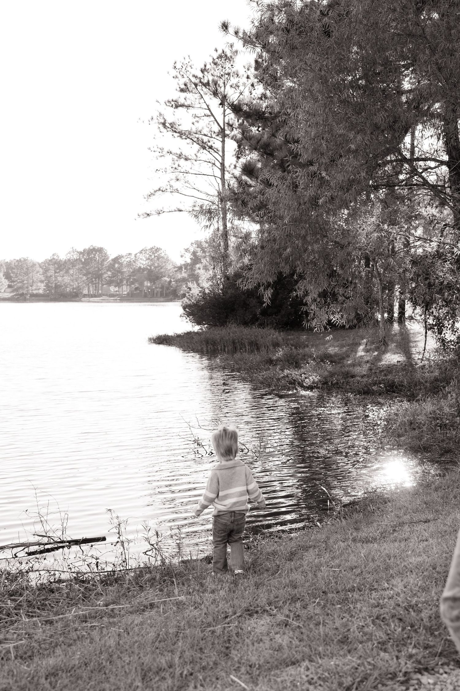 Black and white photo of a young child standing at the edge of a calm lake surrounded by trees.