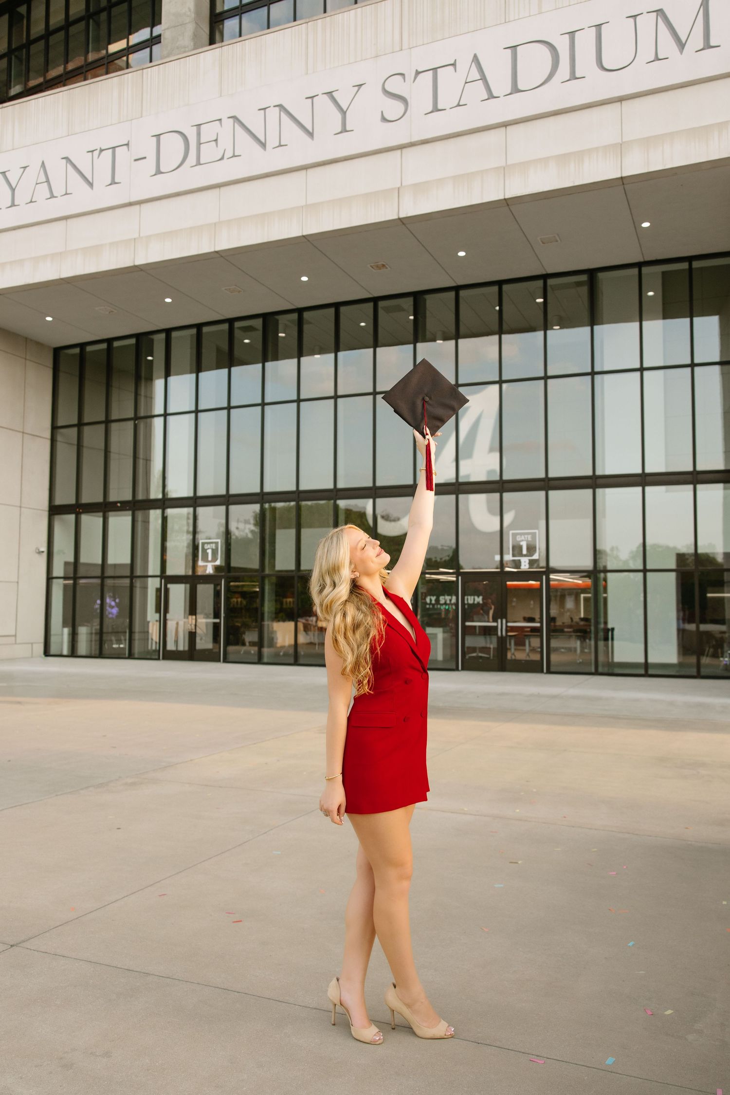 Blonde graduate in red dress raises cap outside Bryant-Denny Stadium at University of Alabama.