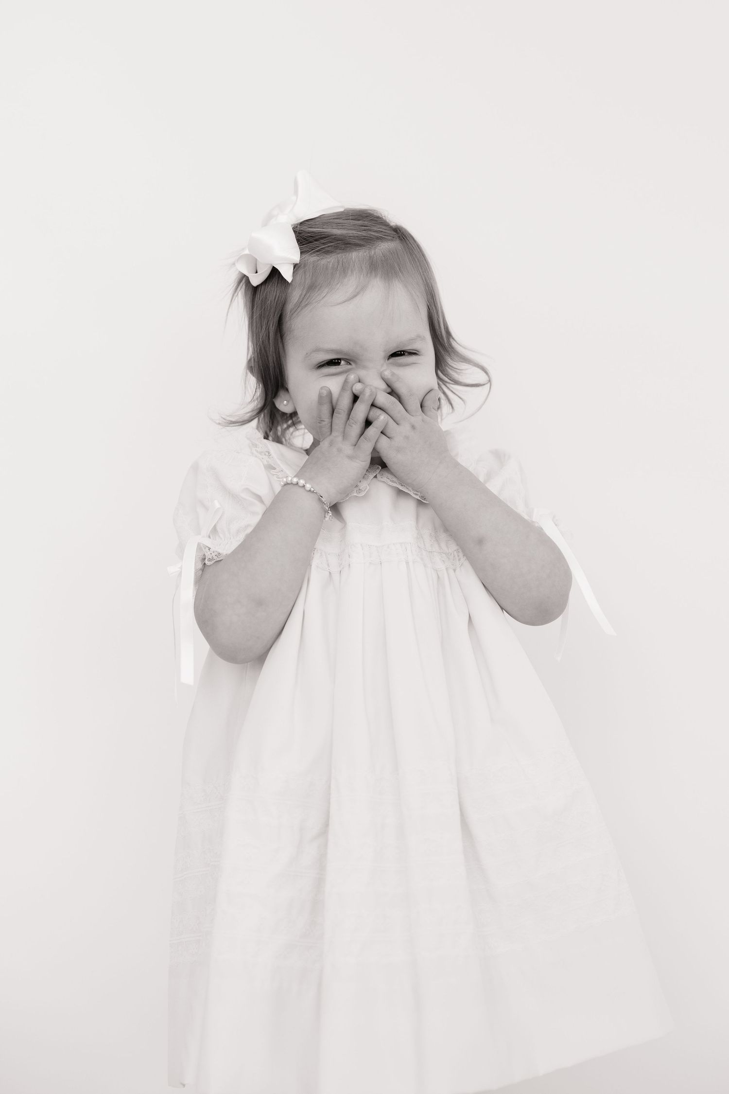 A cute toddler girl in a white dress with bow covers her mouth giggling, black and white portrait photo.