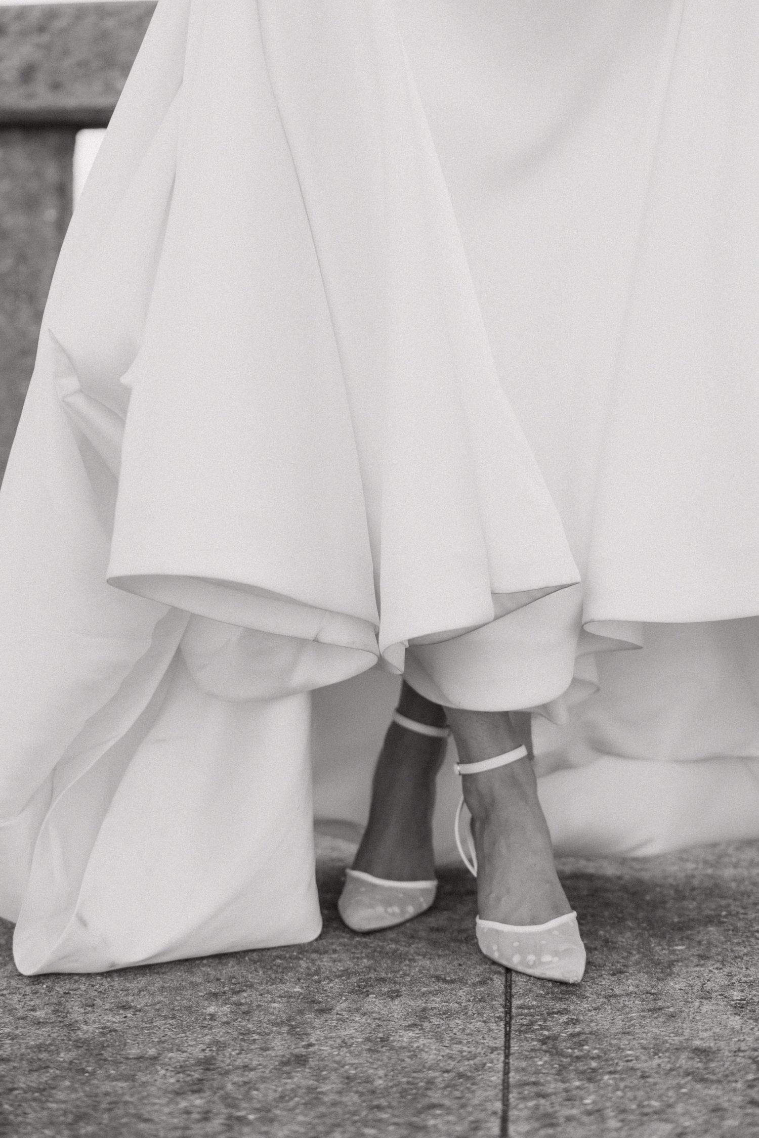 Black and white close-up of bride's white wedding gown hem and elegant ankle-strap heels on stone steps.