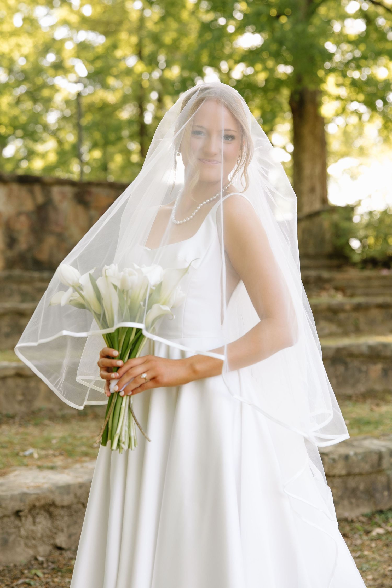 Bride in white gown and veil holding white calla lily bouquet outdoors.