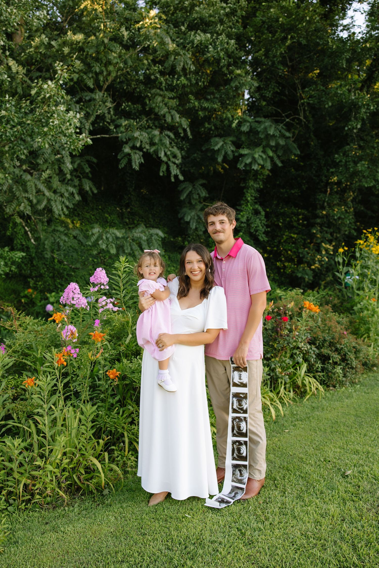 Young family poses in garden, pregnant mother in white dress holds toddler in pink beside husband holding ultrasound photos.