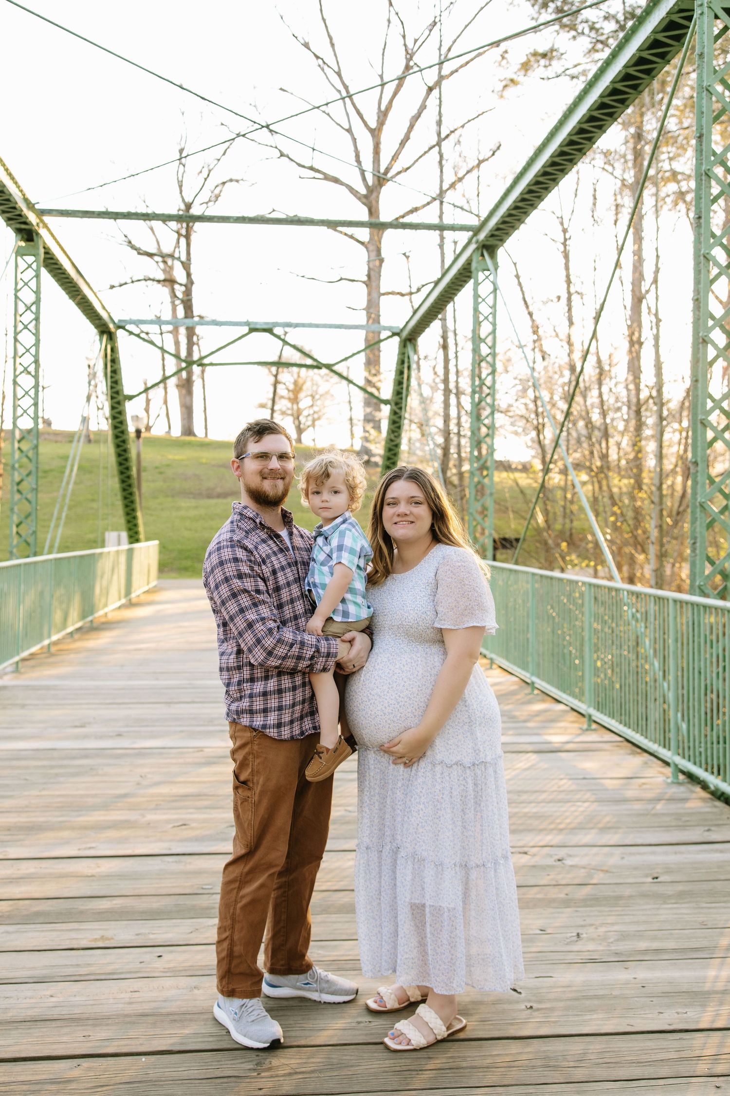 A family maternity photo on a green bridge, with a man holding a toddler beside a pregnant woman in a white dress.