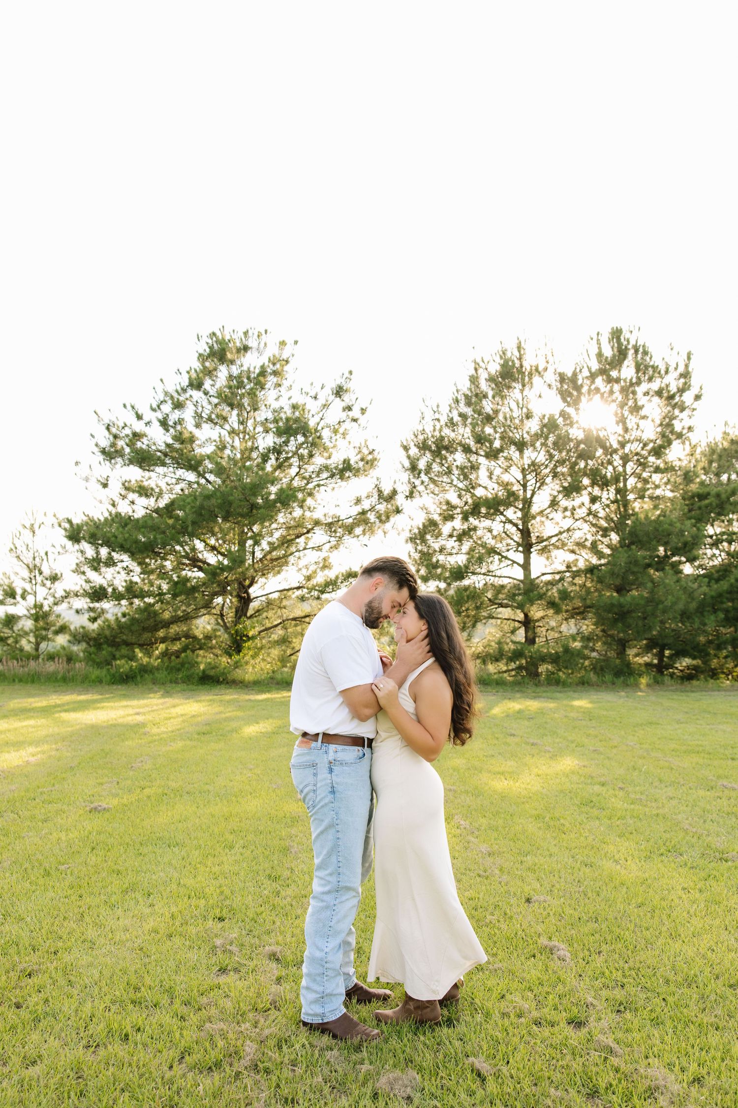 Couple sharing a romantic kiss in a sunlit green field, woman in white dress holding flowers.