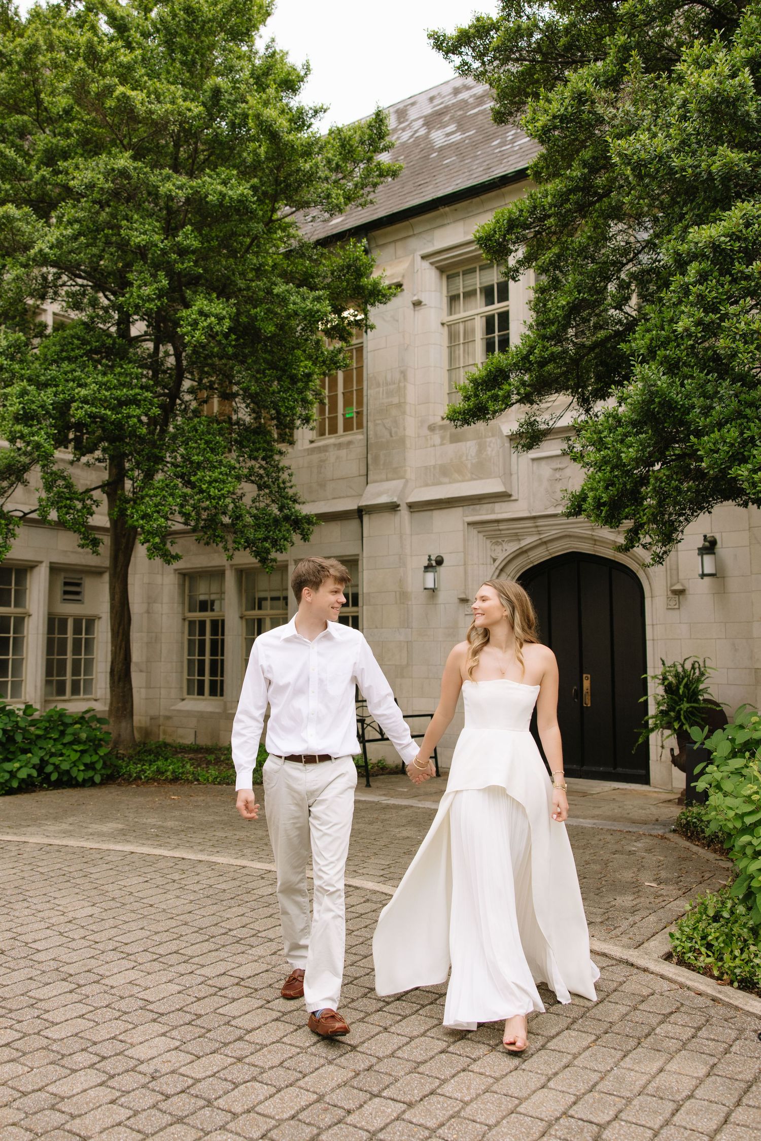 A bride and groom walk hand-in-hand in white attire outside an elegant stone manor surrounded by lush green trees.