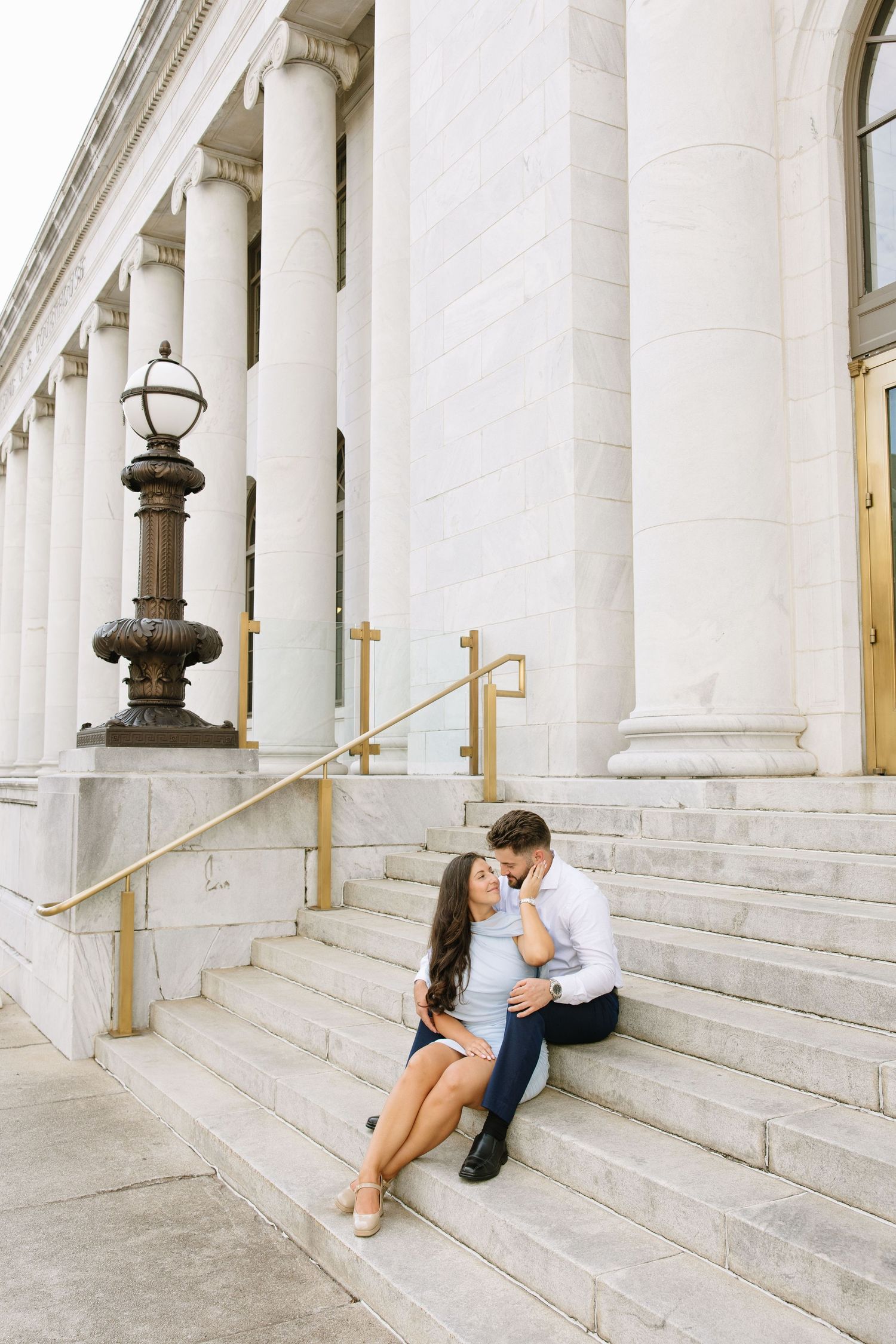 Couple sitting on marble steps of a classical building with white columns, sharing a romantic moment during an engagement session.