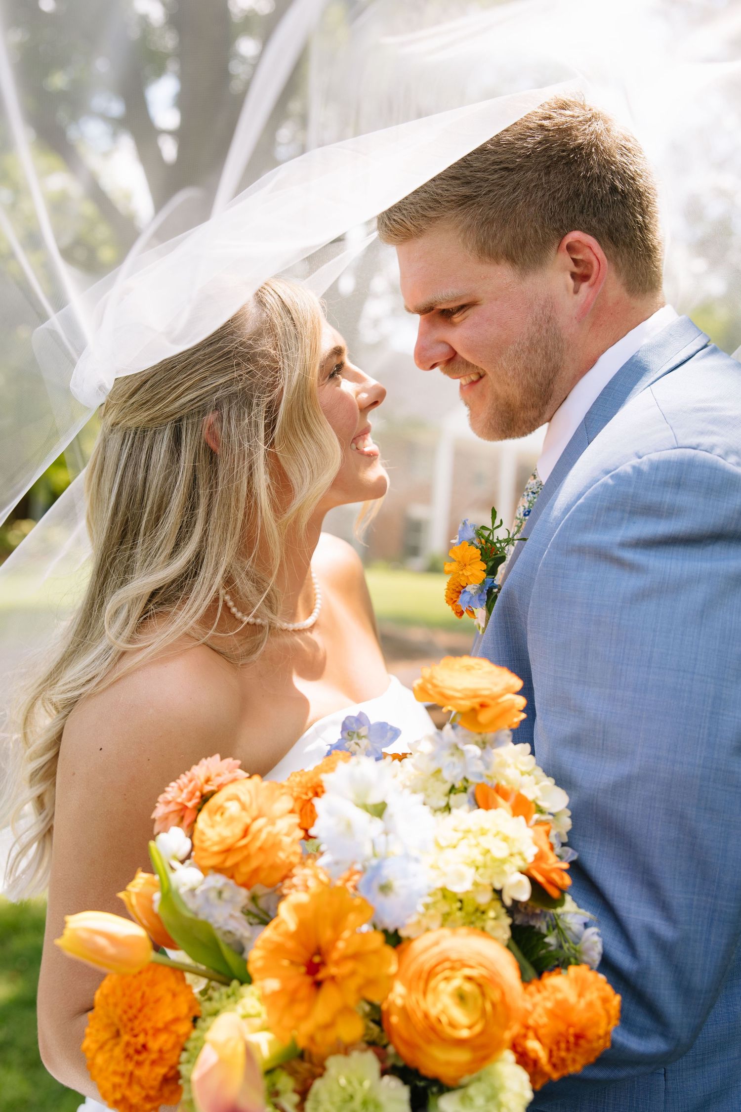 Bride and groom smiling at each other under a veil, holding a vibrant orange and white wedding bouquet outdoors.