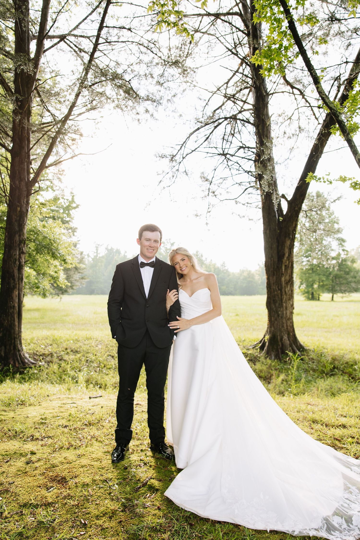 Bride and groom posing together in sunlit green meadow, groom in black tuxedo and bride in strapless white gown.