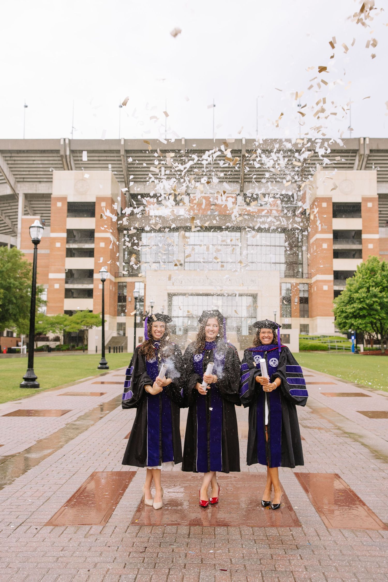 Three graduates in black and purple robes celebrate with confetti outside a stadium on graduation day.