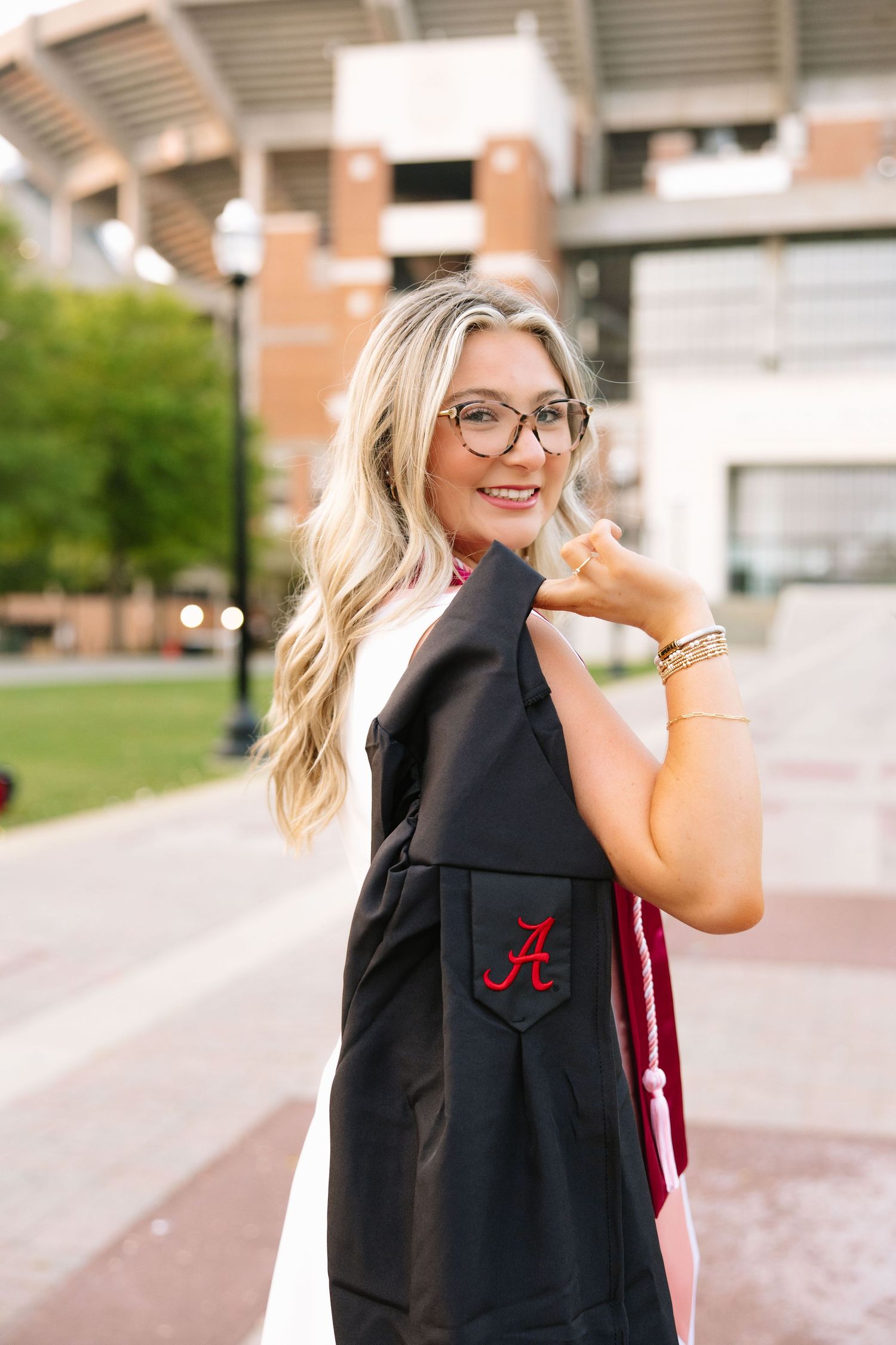 Smiling blonde graduate holds University of Alabama cap and gown outdoors near campus stadium.