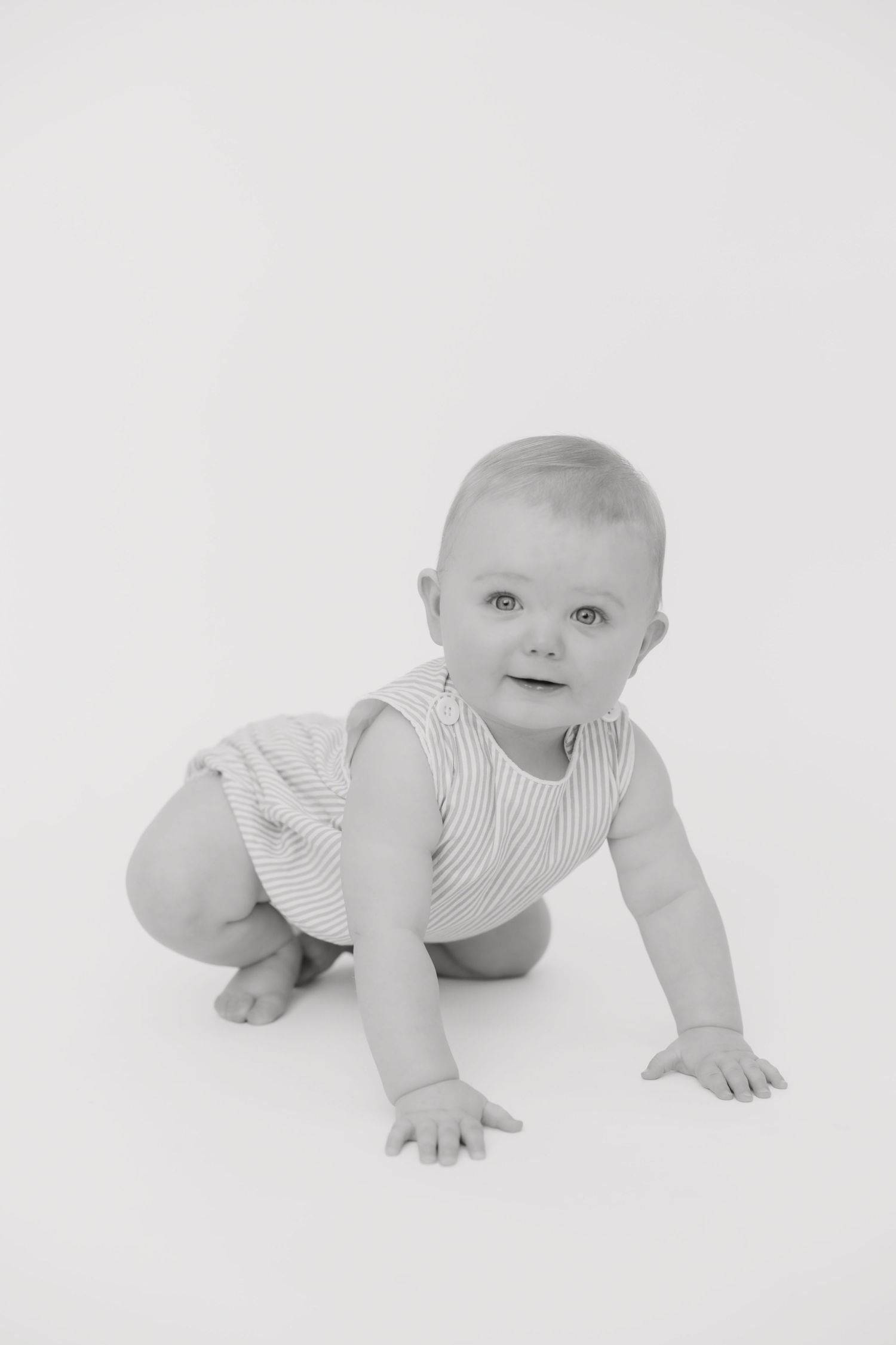 Black and white photo of a cute baby crawling on a white background, wearing a striped romper.