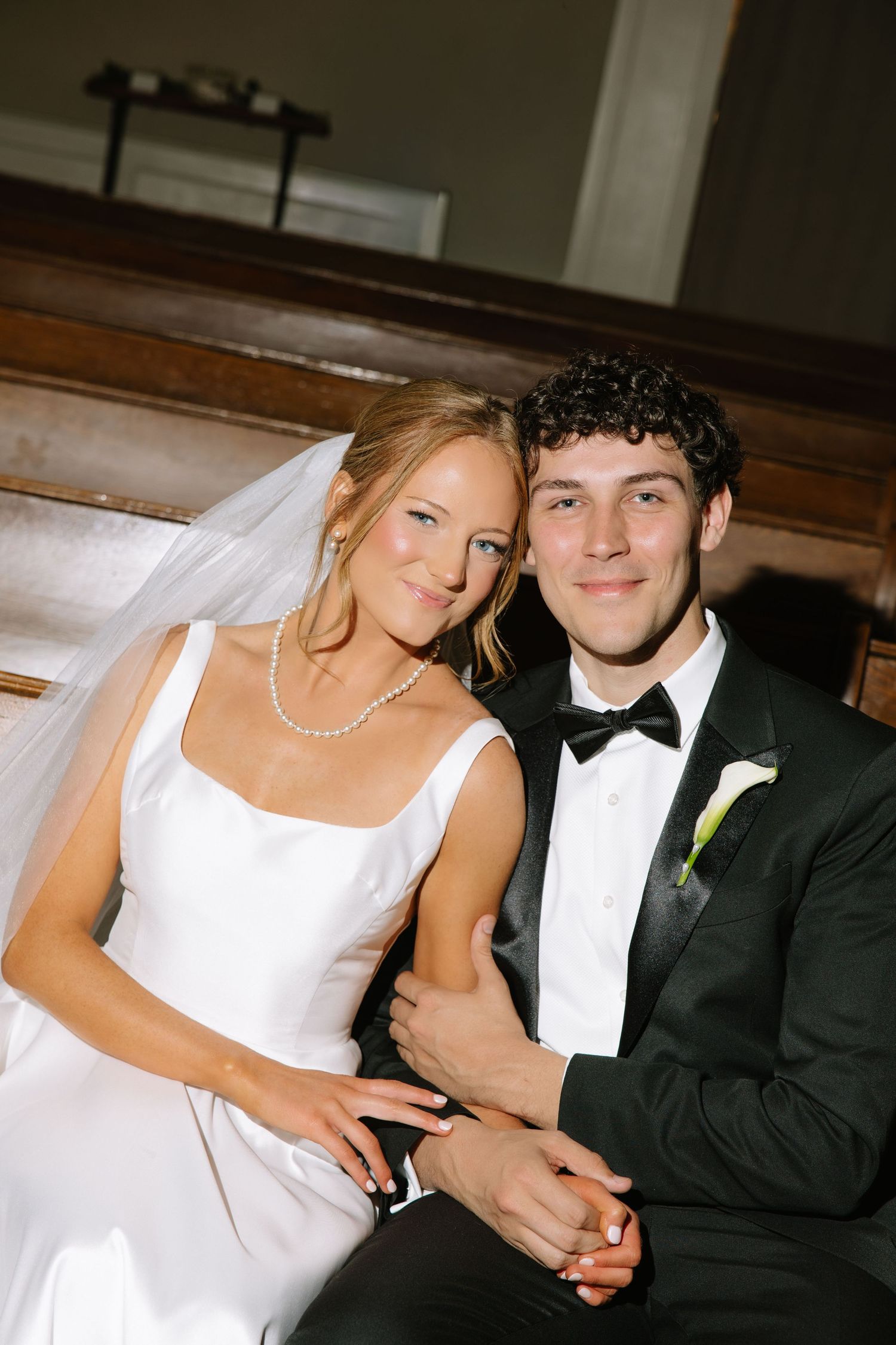 A bride and groom pose together in a church pew, smiling, wearing a white gown and black tuxedo.