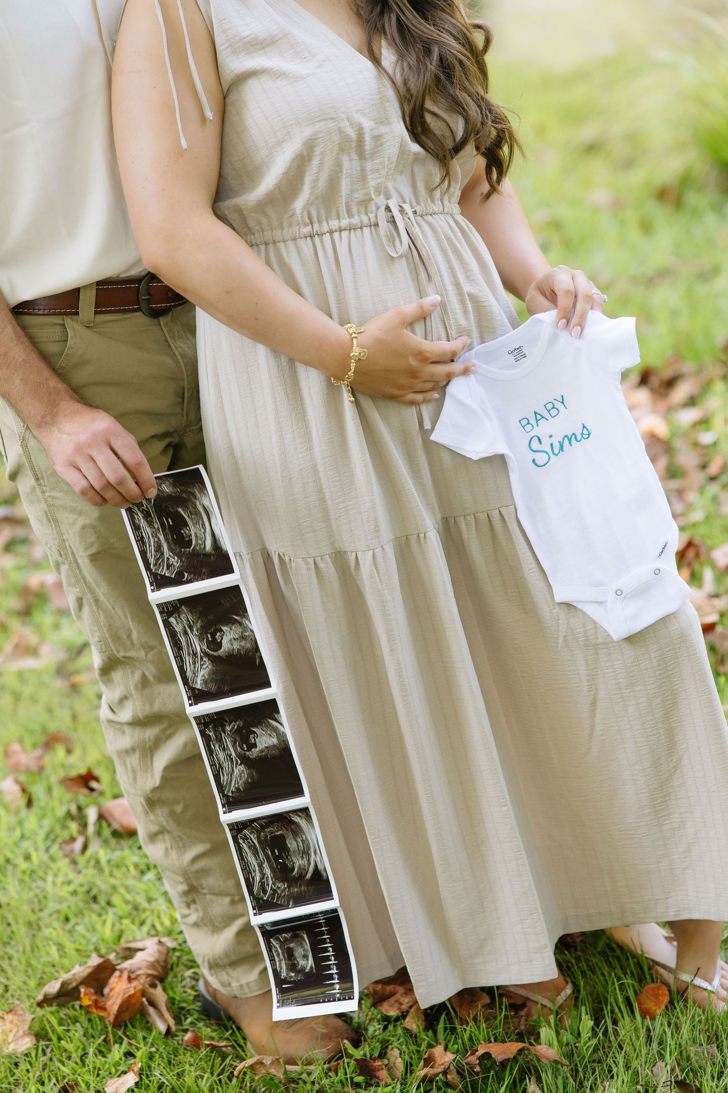 Pregnant couple holding ultrasound photos and a white Baby Sims onesie outdoors on green grass.
