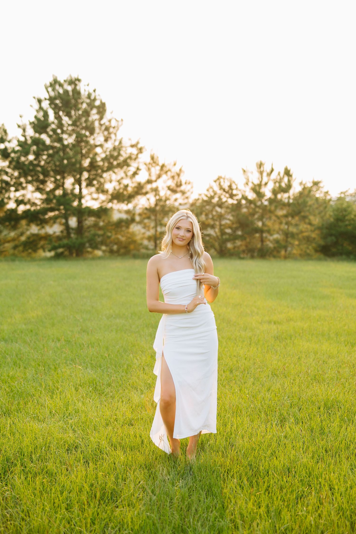 Young woman in white strapless dress standing in green field at golden hour with pine trees in background.