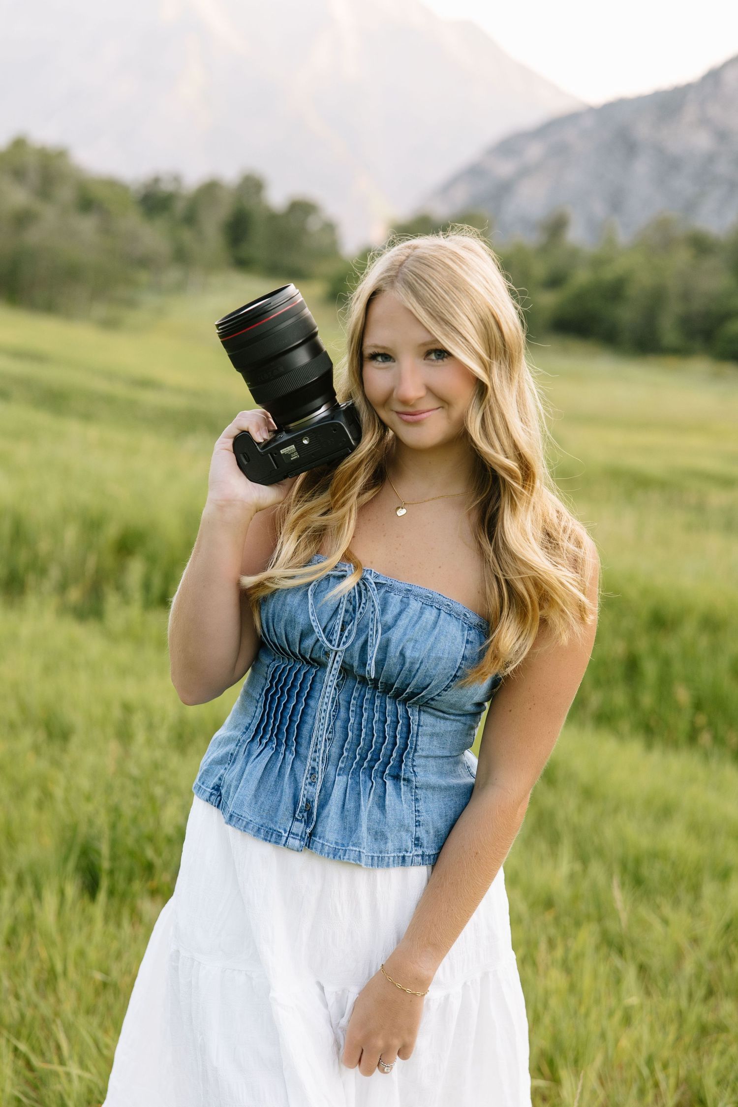 Young blonde woman holding a DSLR camera in a green mountain meadow, wearing a denim top and white skirt.