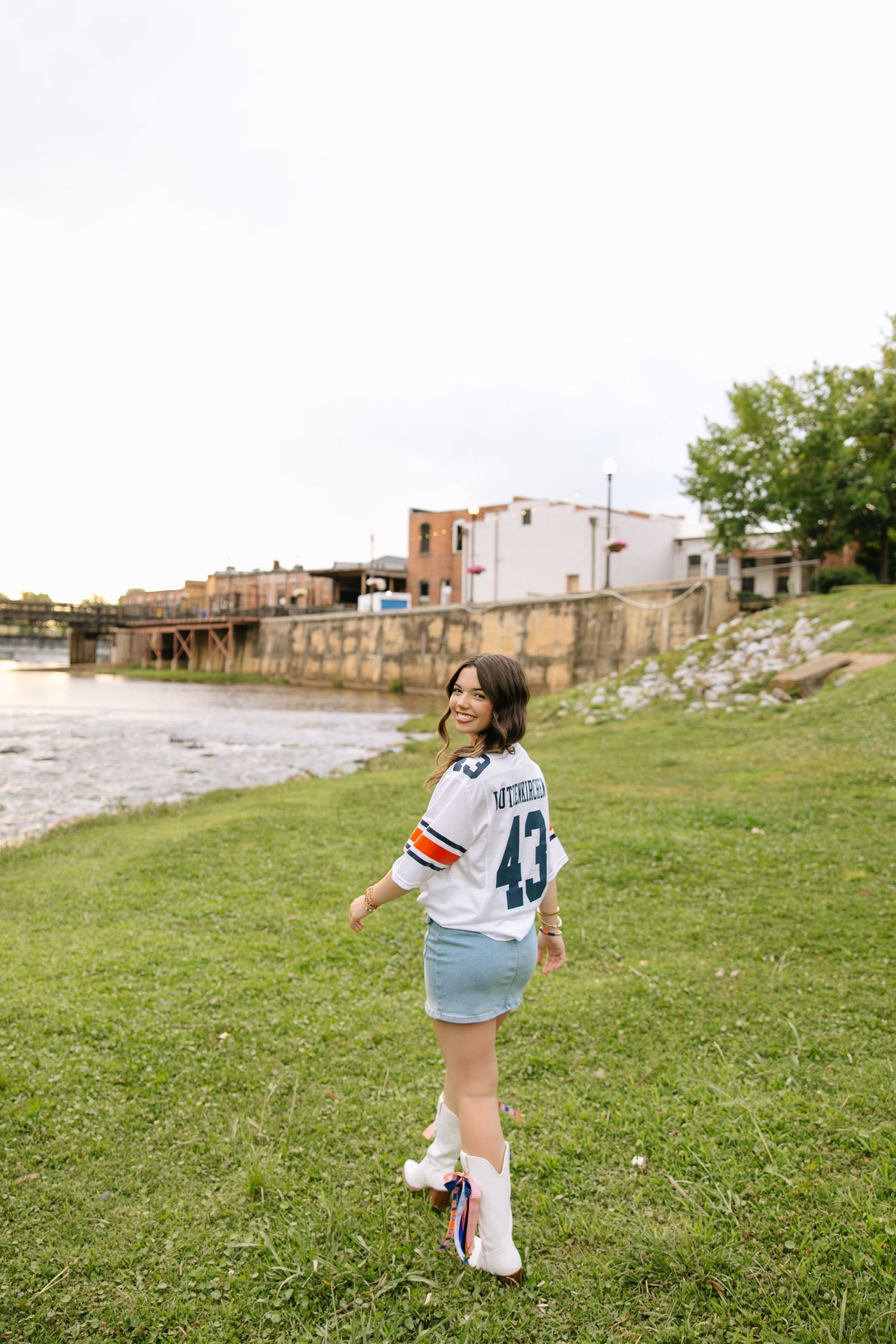 Young woman in football jersey and denim skirt poses by riverside with historic buildings in background.