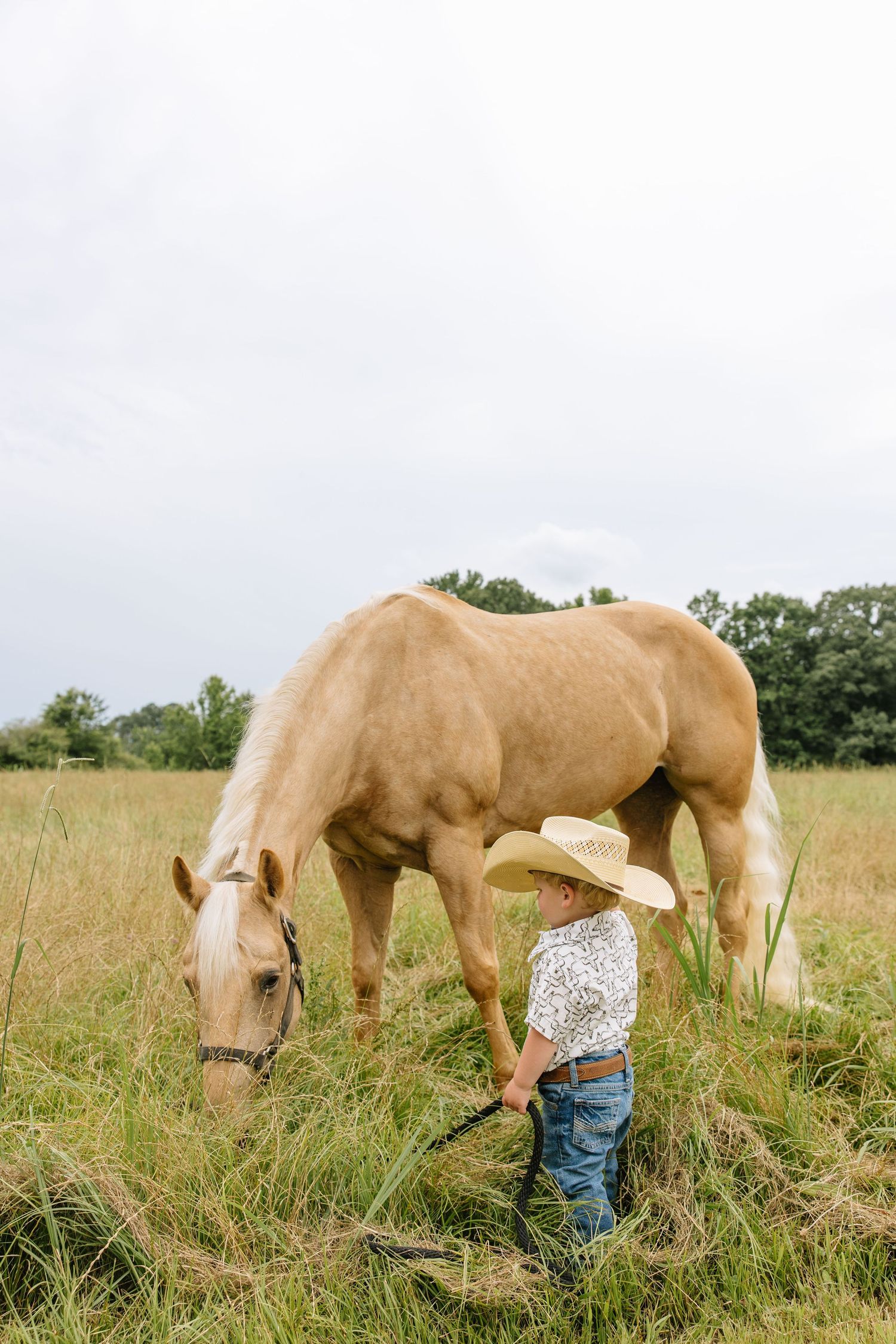 Young child in cowboy hat standing beside a palomino horse grazing in a green field.