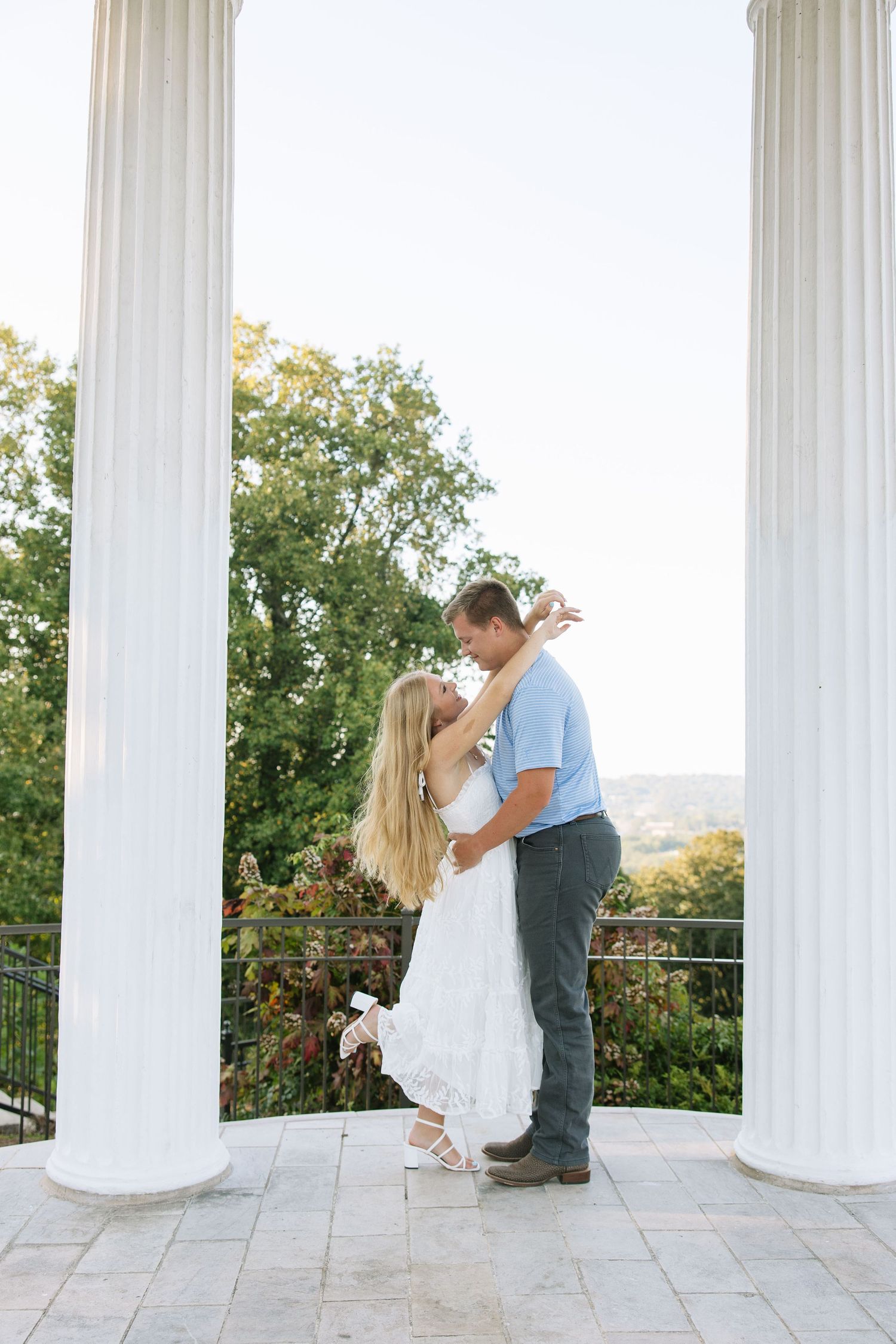 Couple sharing a romantic kiss between white columns on a scenic outdoor terrace with lush green trees in the background.