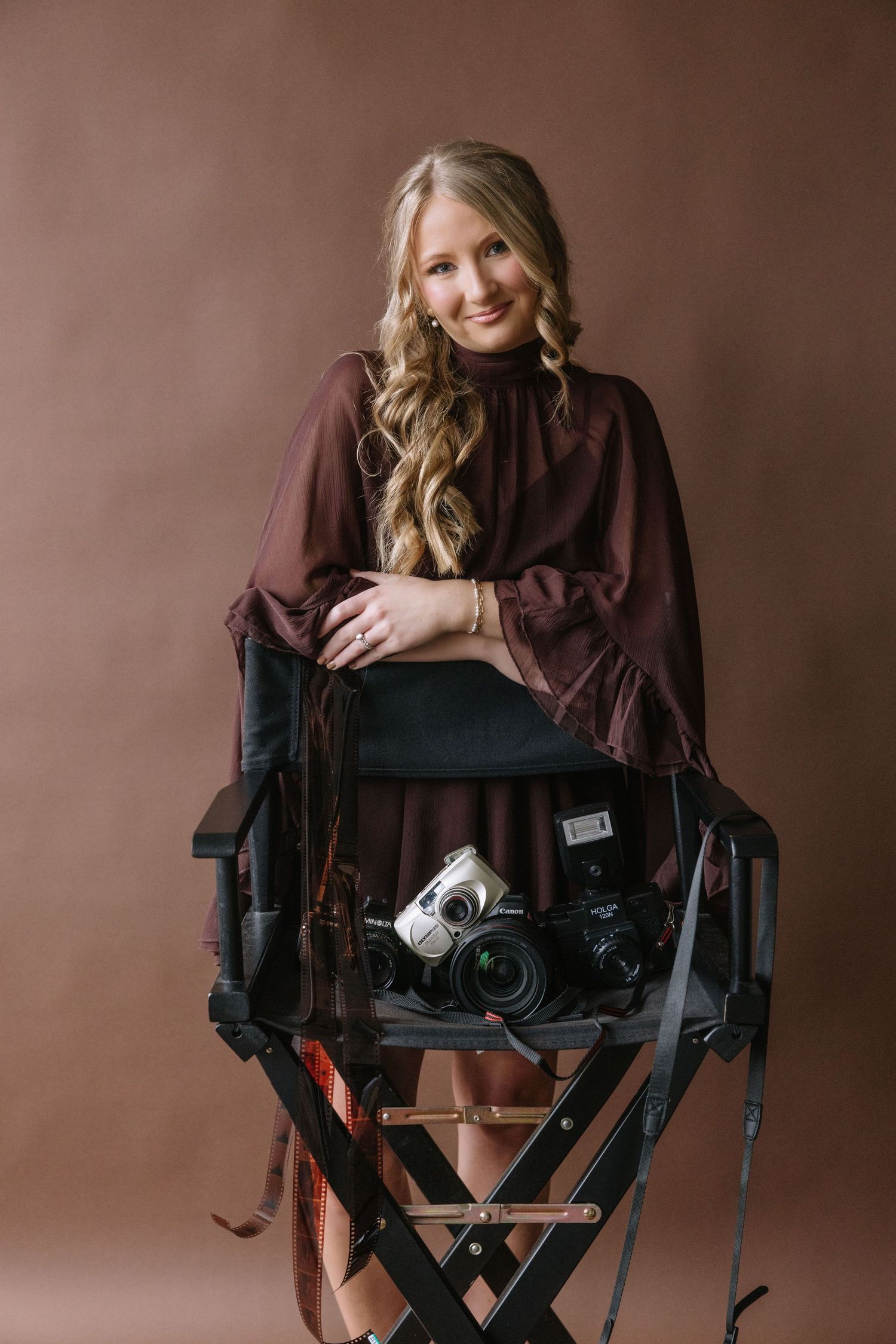 Smiling blonde woman in burgundy blouse sitting on director's chair with cameras, brown studio backdrop.