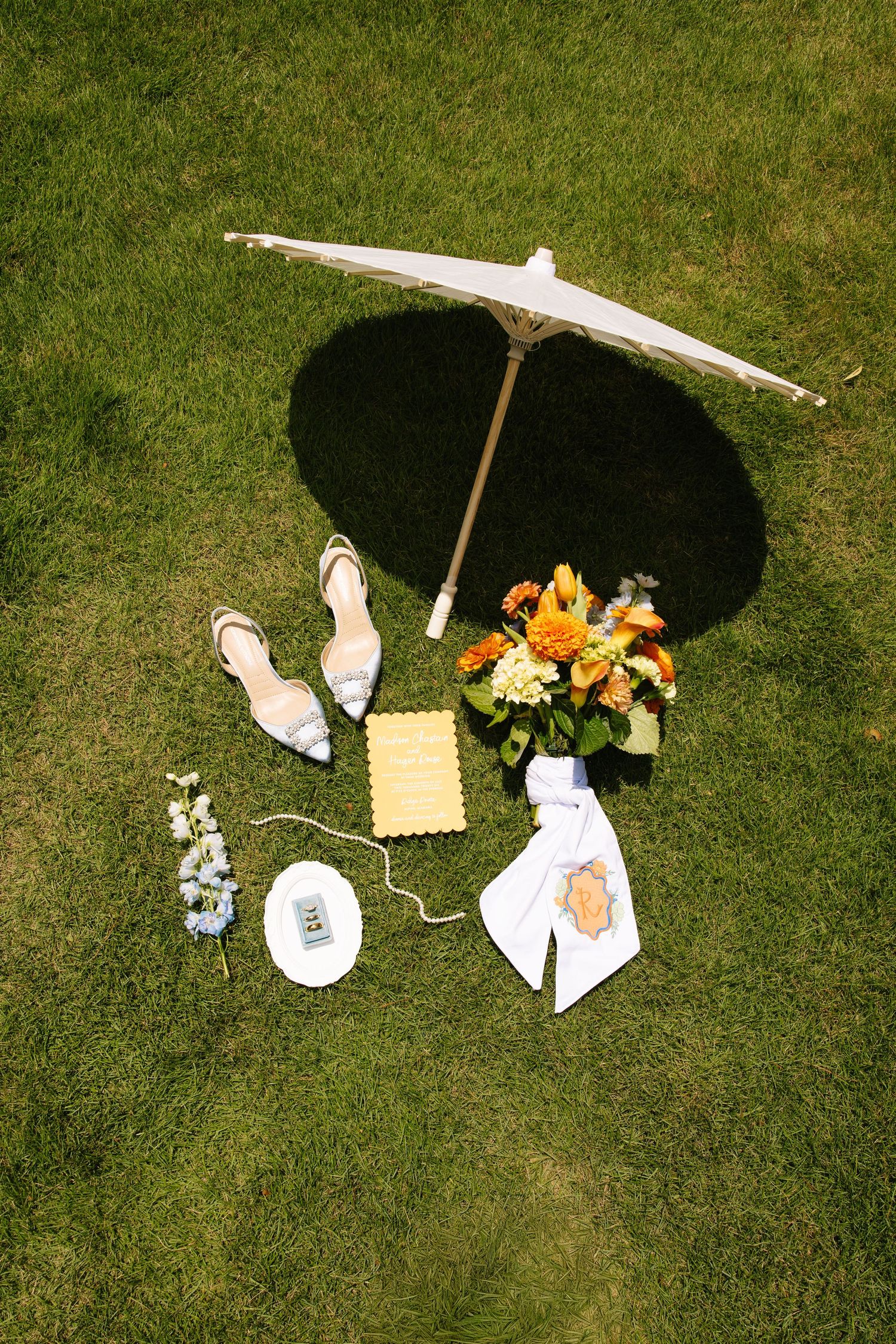 White parasol, bridal heels, orange floral bouquet, and wedding accessories arranged on lush green grass.